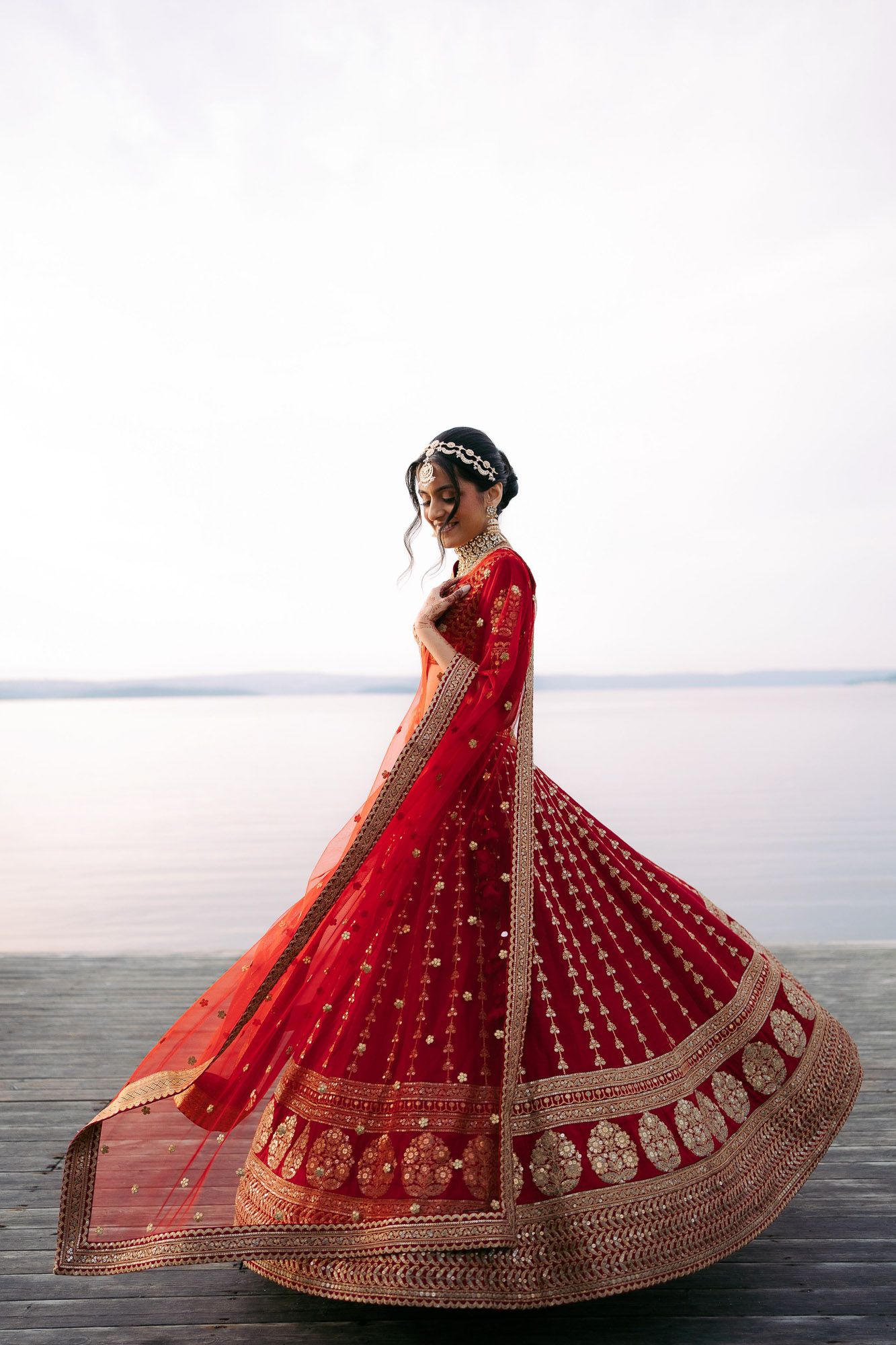 A bride in a stunning red and gold embellished lehenga twirls gracefully against an ocean backdrop.