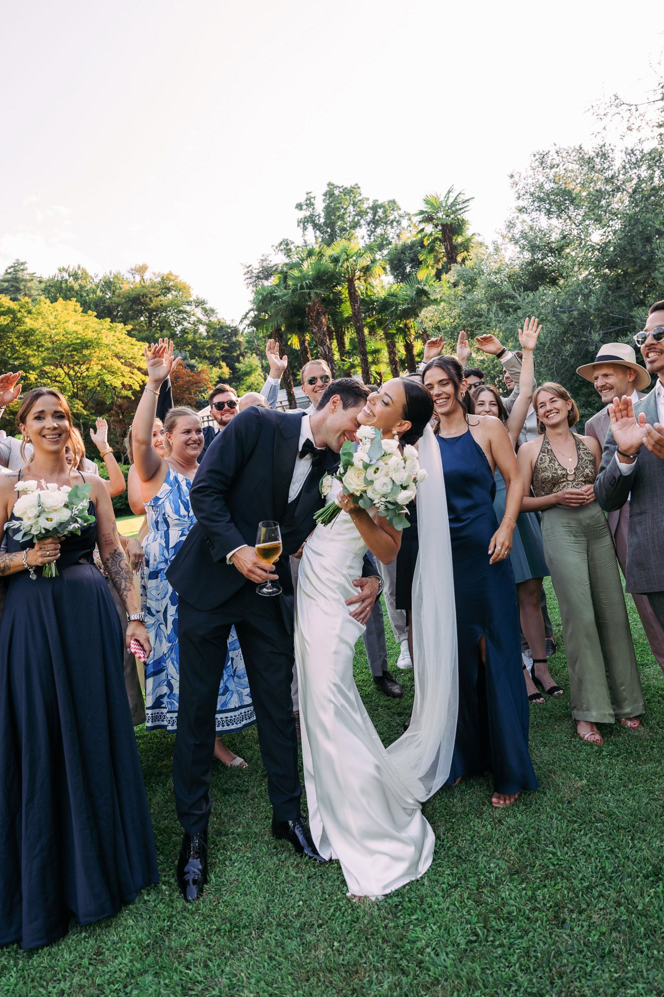 Wedding celebration on a lush green lawn with bridesmaids in navy dresses and guests cheering the newlyweds.