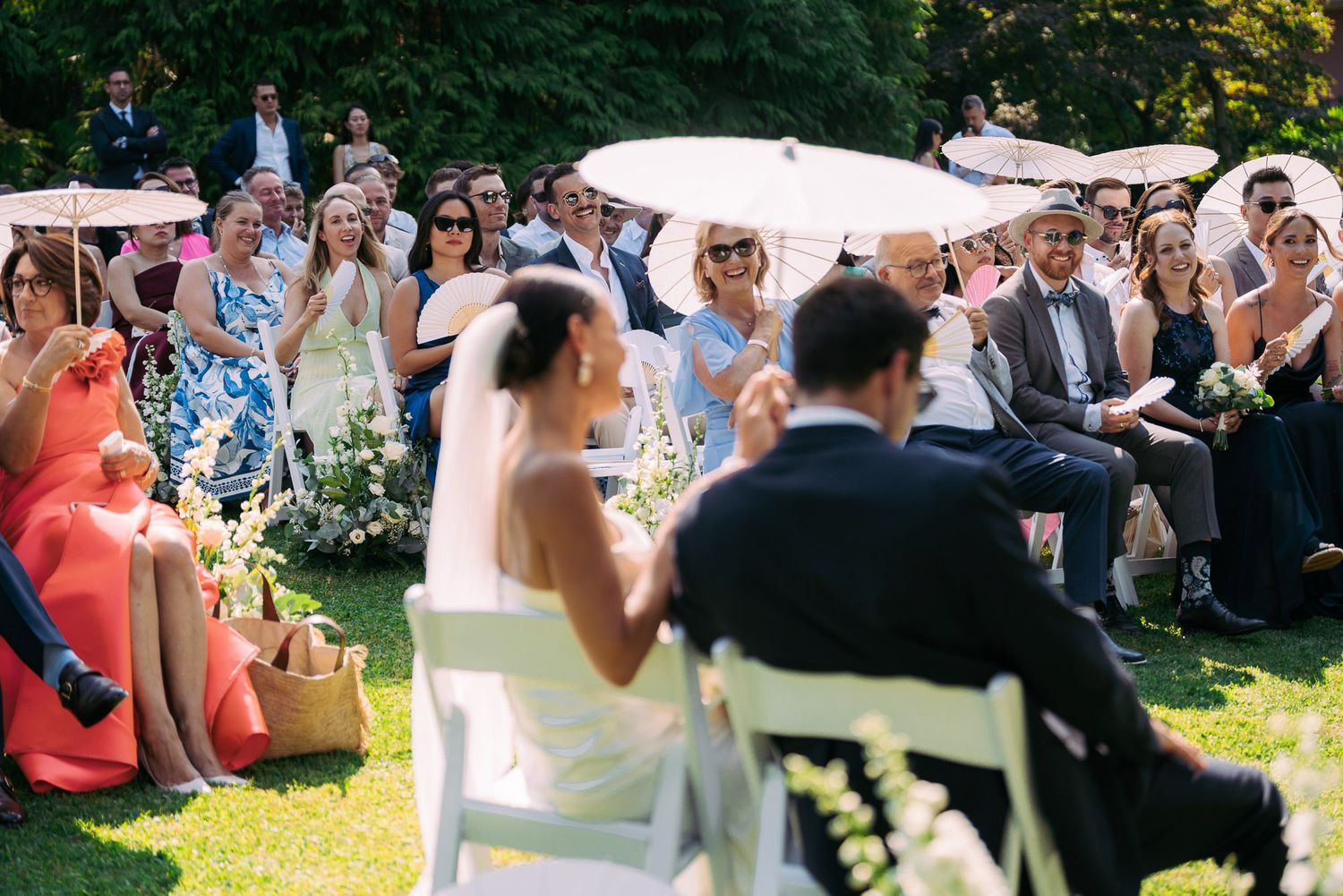A group of wedding guests sit outdoors on white chairs during a sunny garden ceremony.