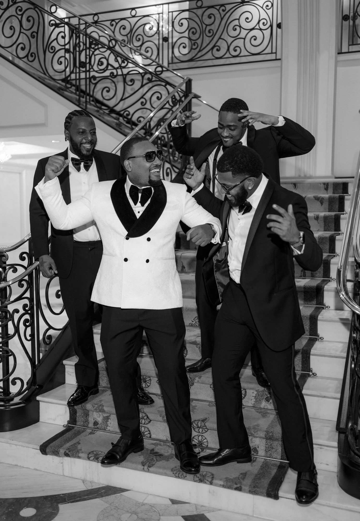 A group of men in formal tuxedos pose playfully on elegant marble stairs with ornate wrought iron railings.