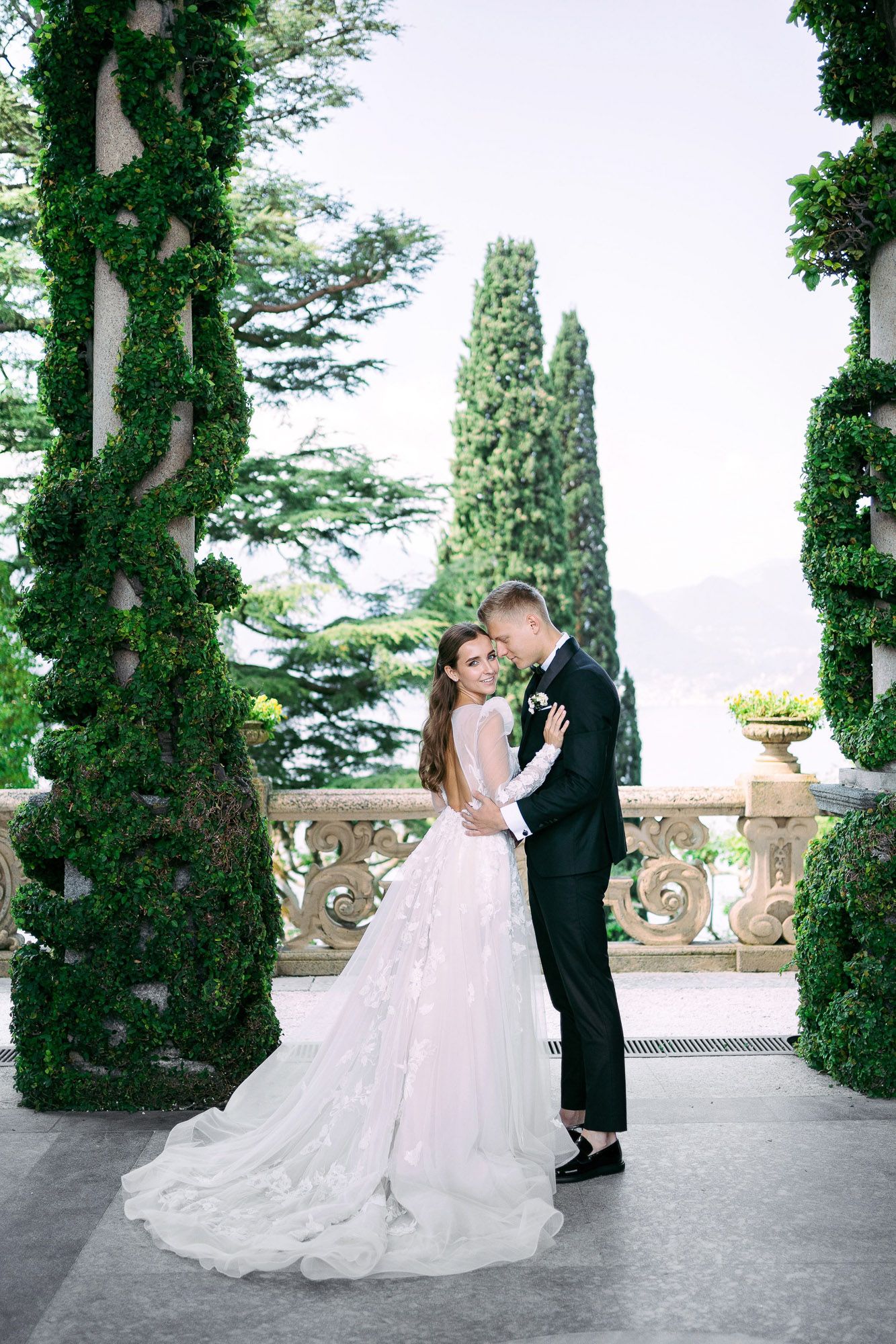 A romantic wedding couple embraces on an elegant stone terrace surrounded by tall cypress trees and classic architecture.