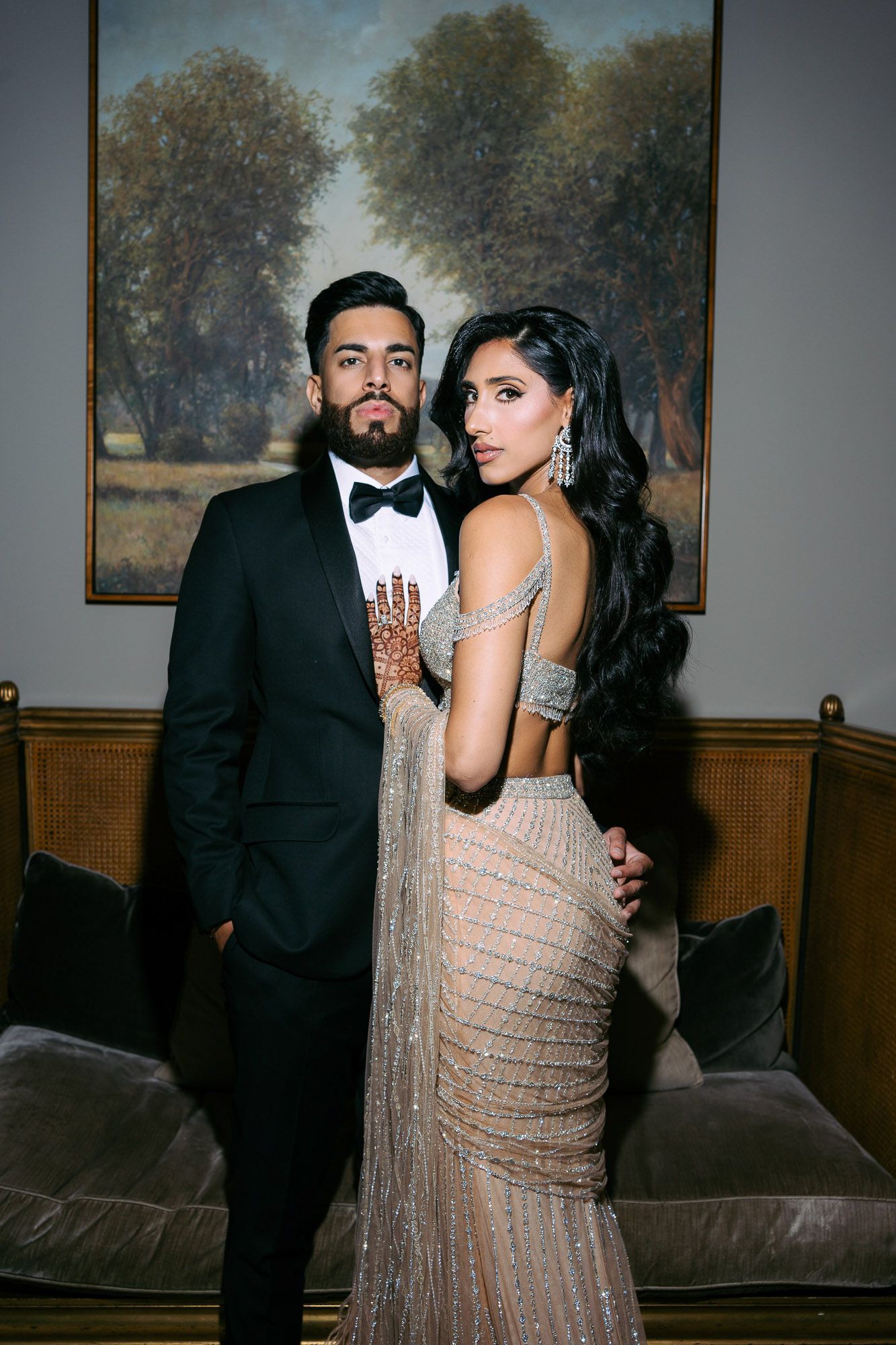 Glamorous couple poses in formal attire at an elegant venue with a landscape painting in the background.