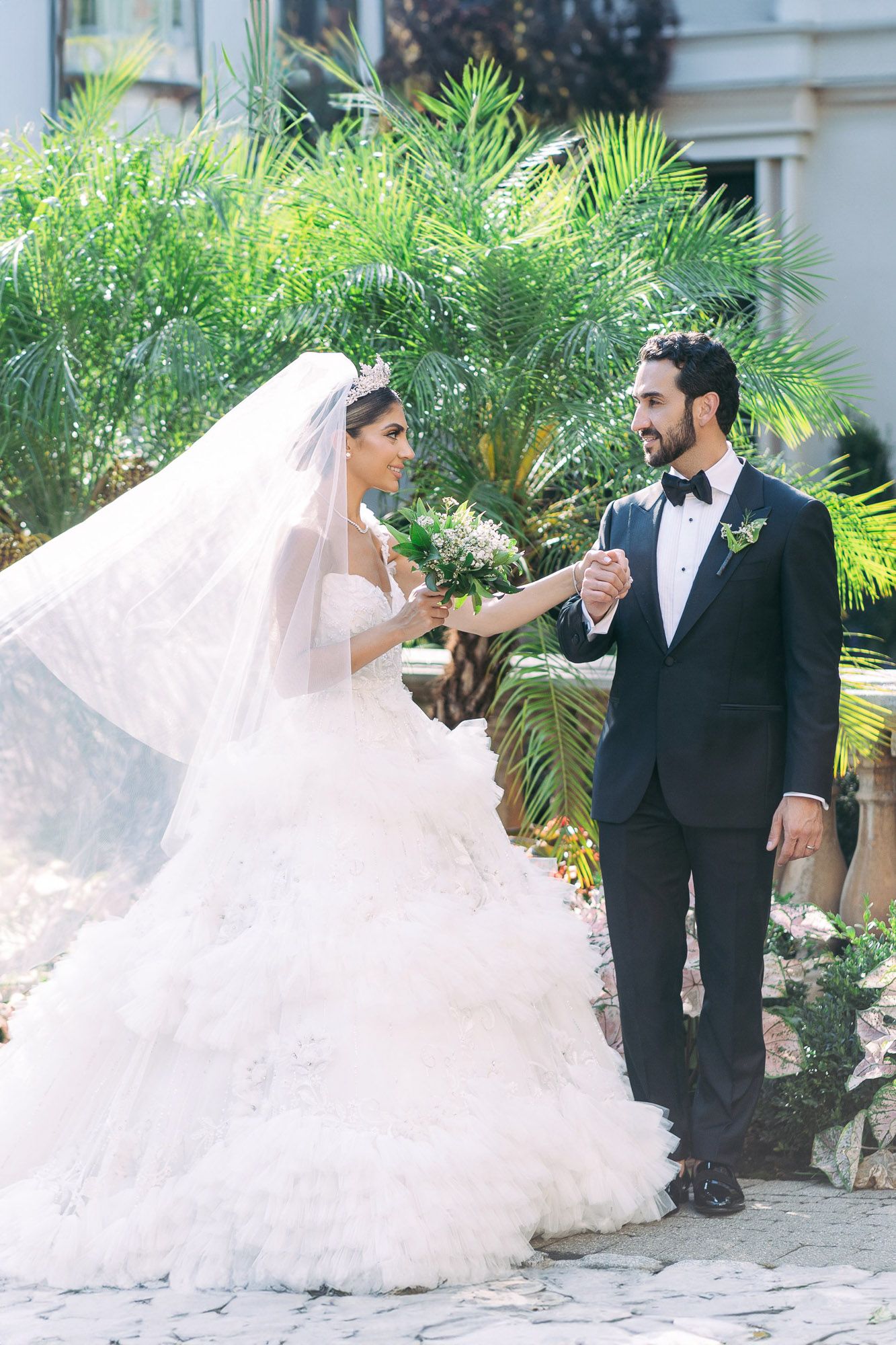 A bride and groom share a romantic moment in a garden setting with flowing white wedding dress and palm trees.