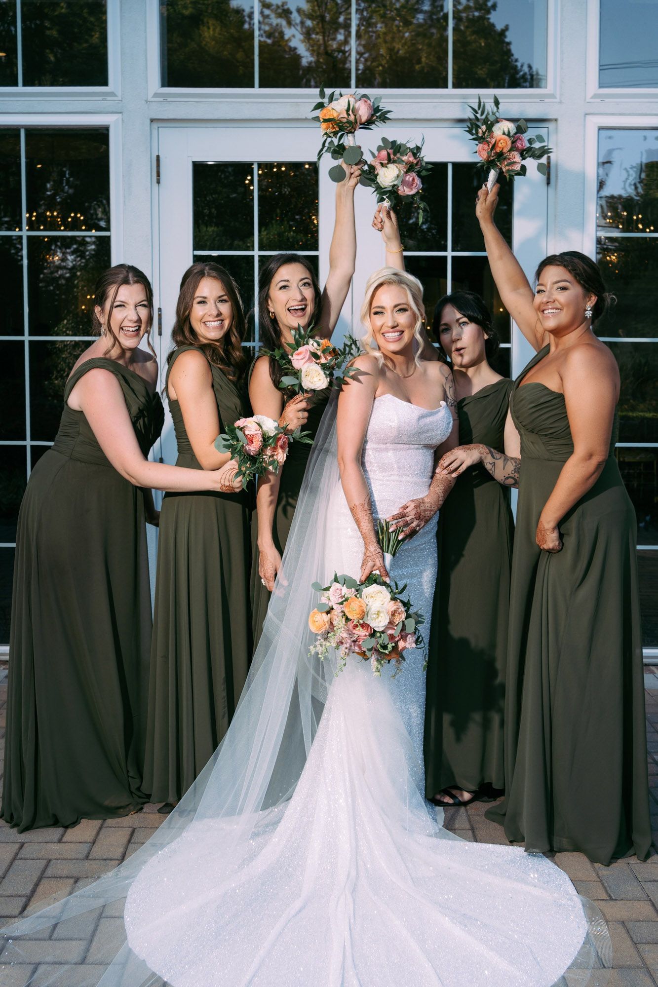 Bridal party in olive green dresses and white gown pose together in front of large windows during wedding celebration.