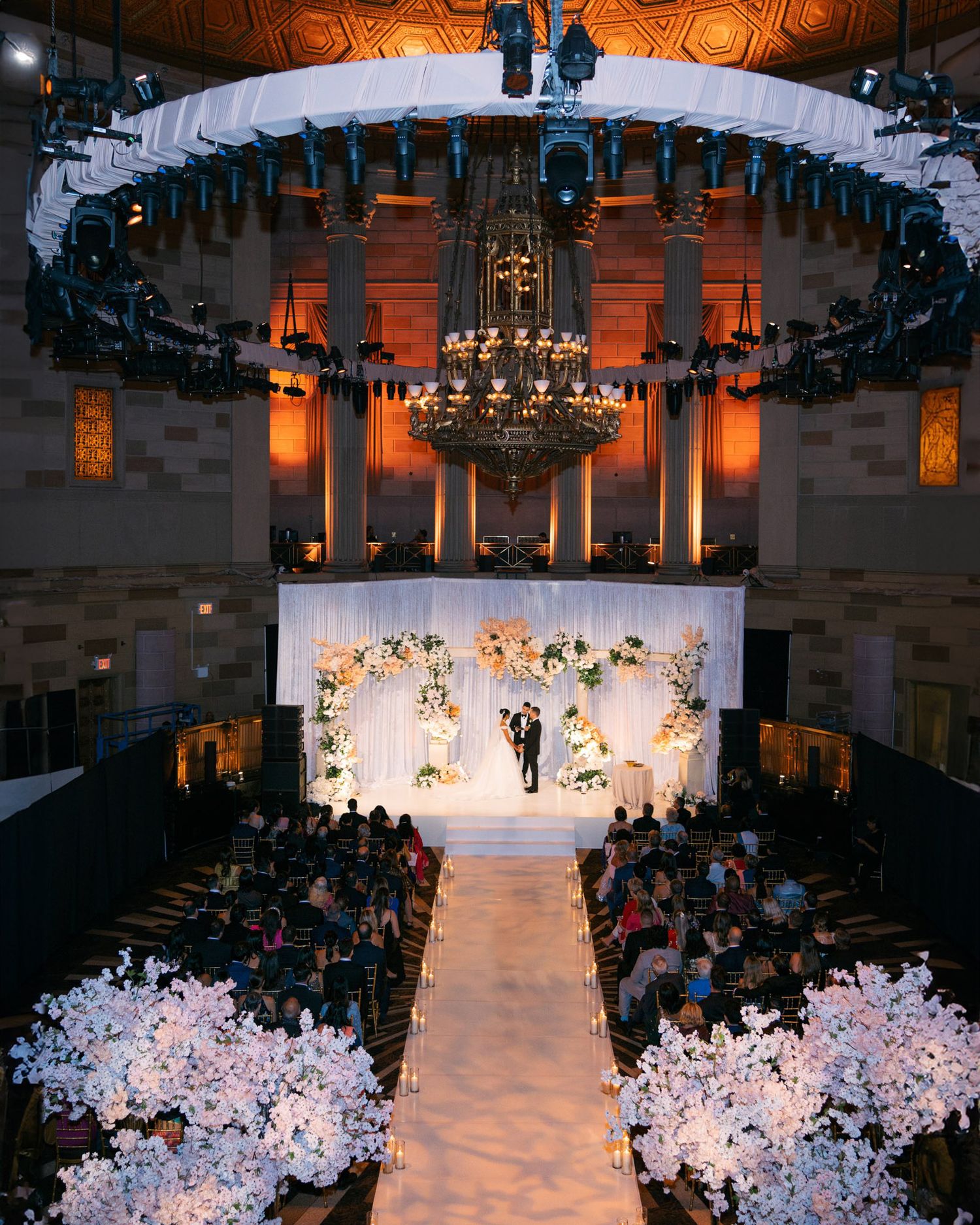 Elegant wedding ceremony setup with white floral arrangements lining the aisle and guests seated in a grand ballroom.