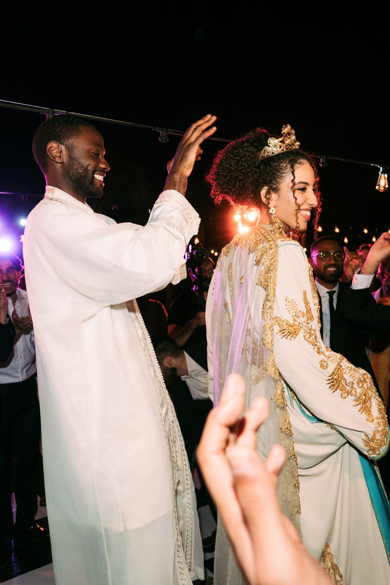 Wedding celebration with couple in traditional white and gold attire dancing under string lights.