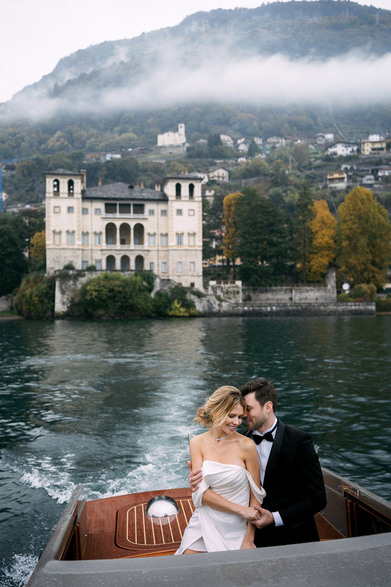 Elegant lakeside villa in Lake Como with misty mountains and autumn trees creates romantic backdrop for boat ride.