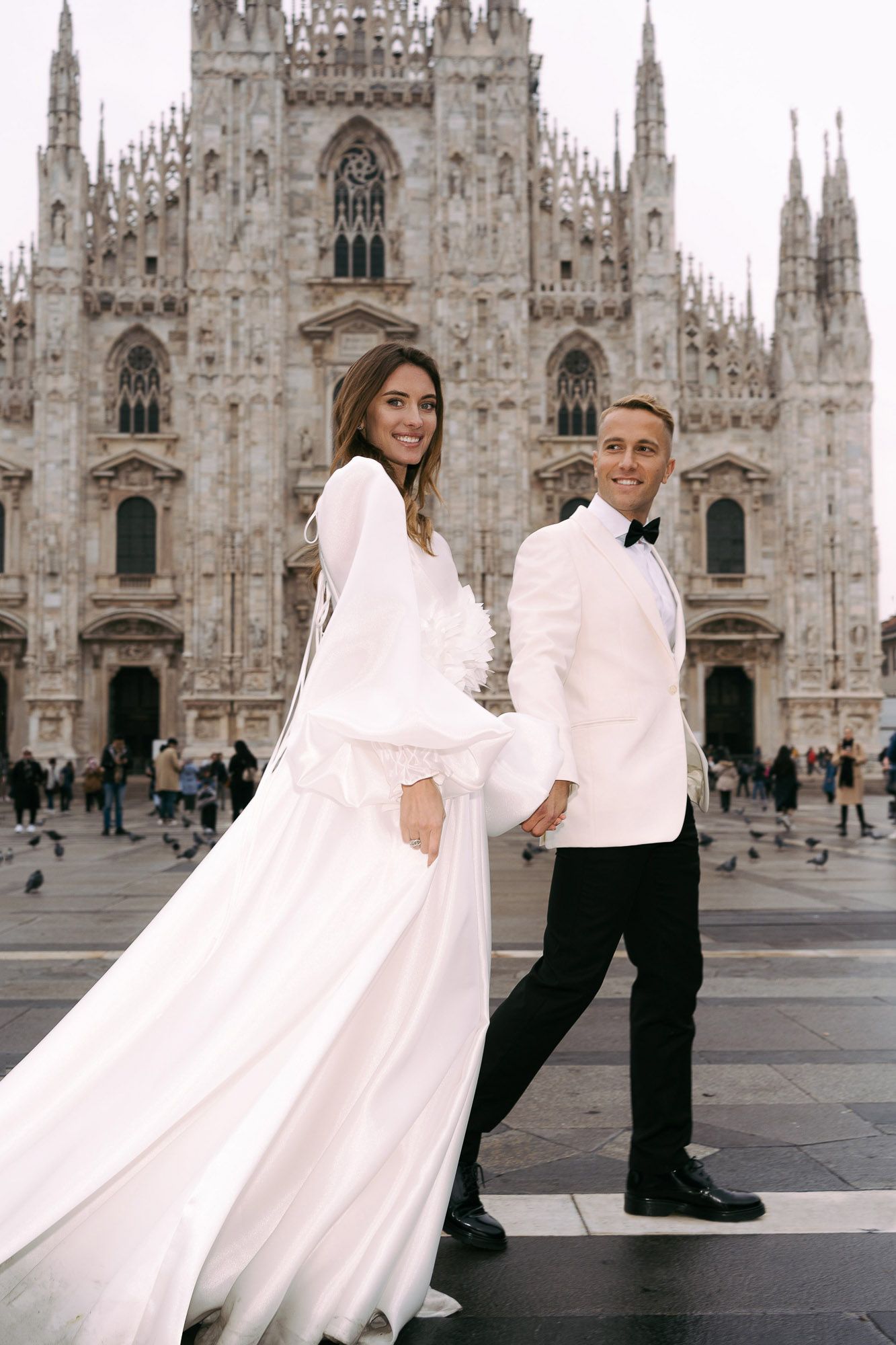 A couple poses in front of Milan Cathedral in Italy, with the bride wearing a flowing white dress against gothic architecture.