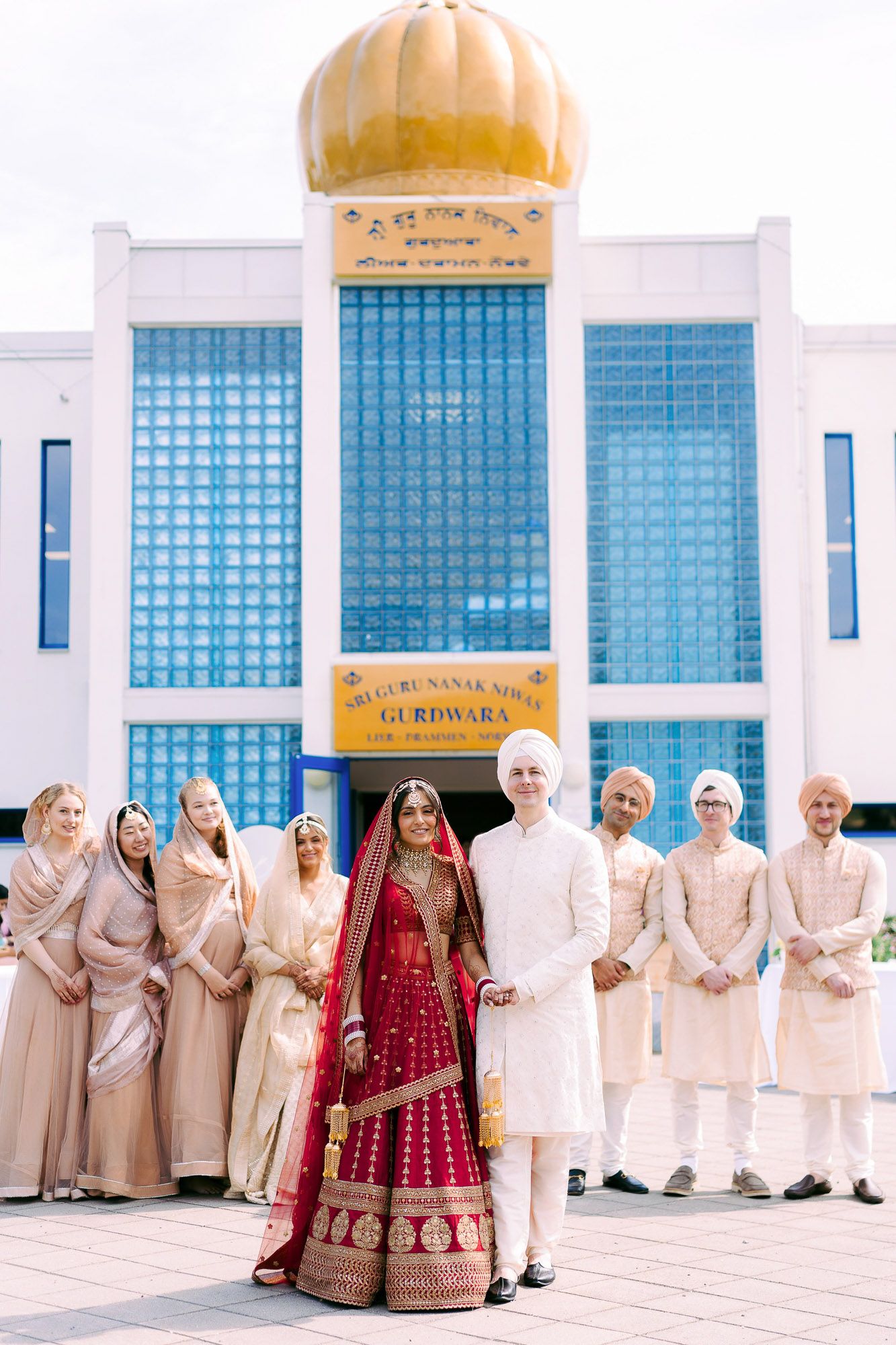 Wedding party poses in traditional Indian and Western attire outside a building with blue glass panels and golden dome.
