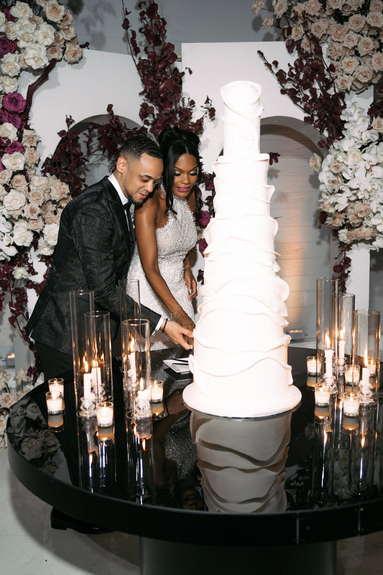 A wedding couple cuts their elegant white tiered cake under an arch decorated with white flowers and glowing candles.