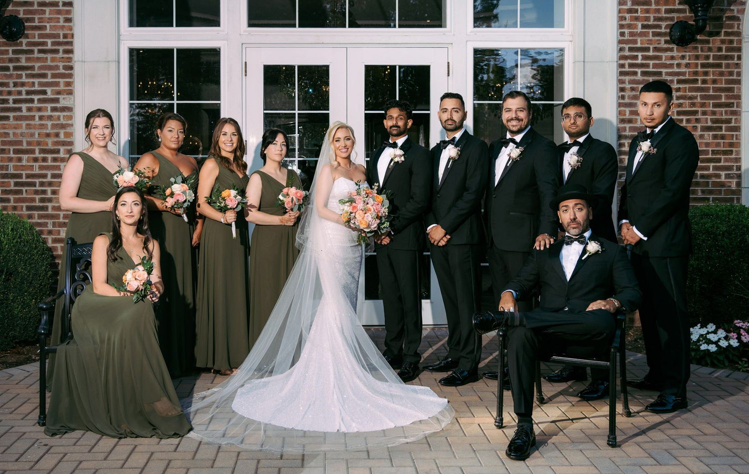 Wedding party poses in formal attire outside brick venue with bridesmaids in olive dresses and groomsmen in black tuxedos.