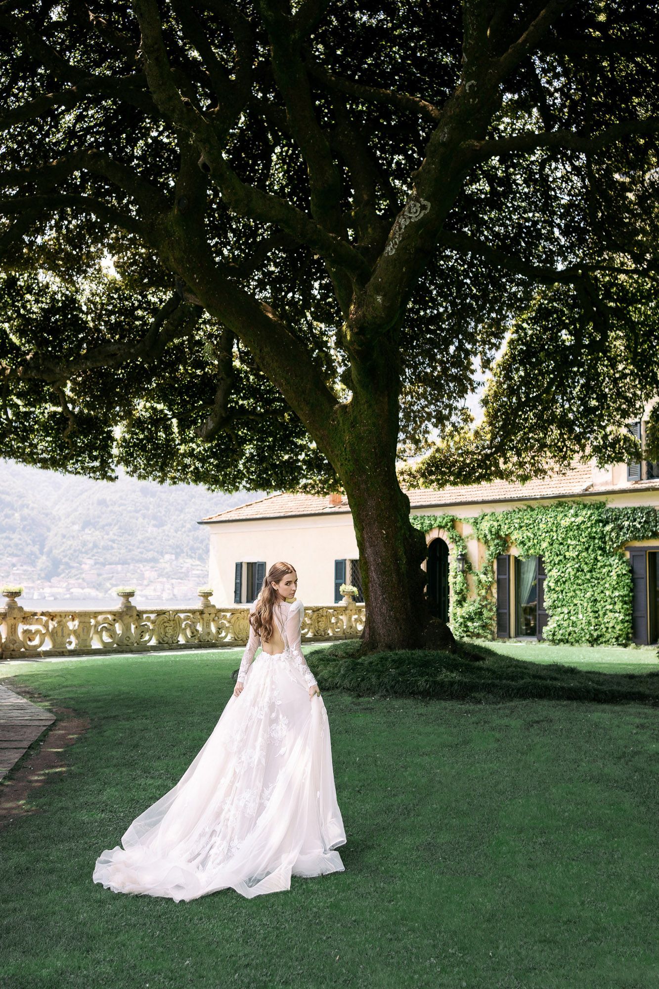 A figure in a flowing white dress spins gracefully on a sunlit lawn surrounded by mature trees and stone buildings.