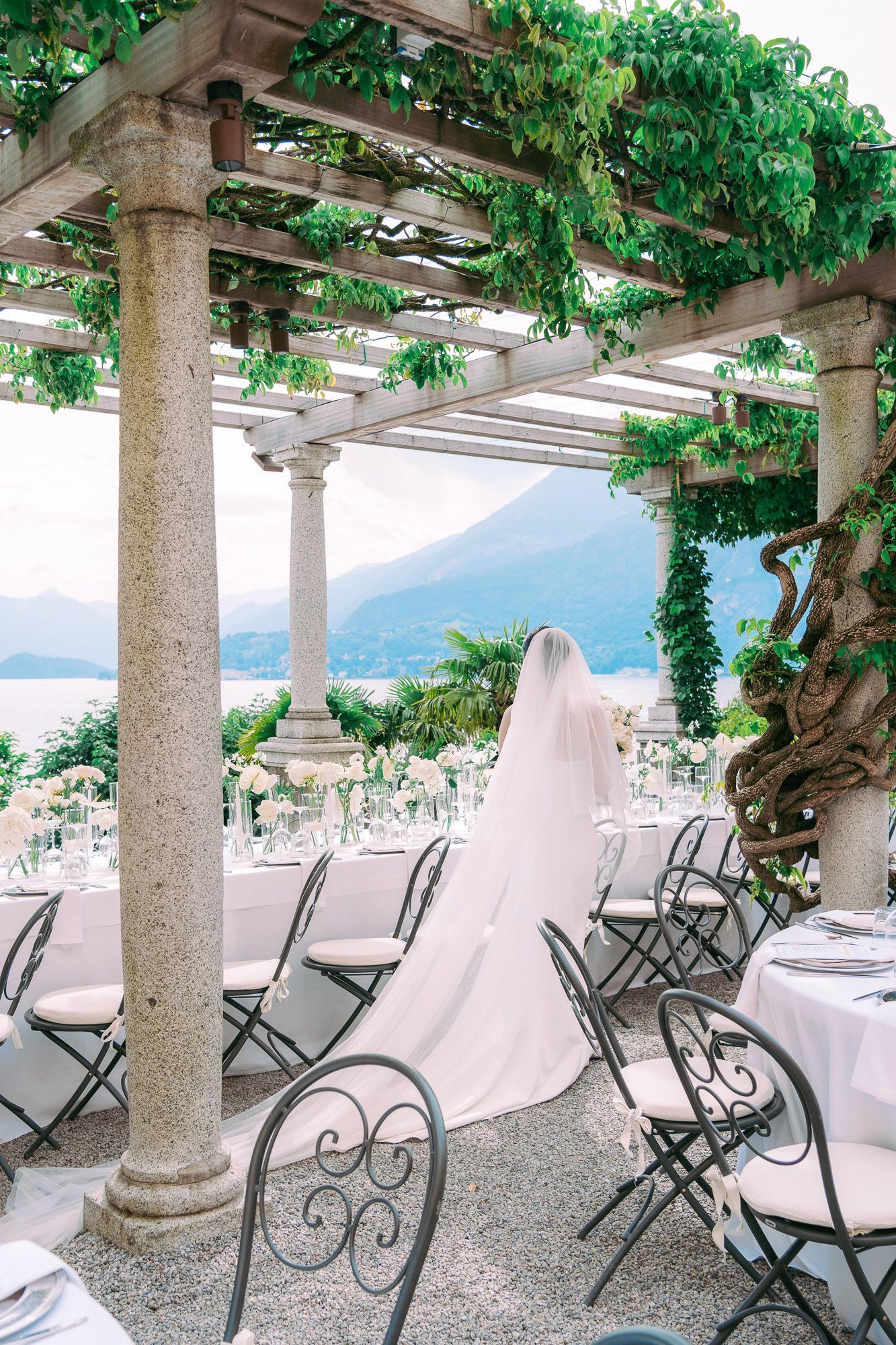 Elegant outdoor terrace with stone columns and climbing vines overlooking a lake and mountains in Italy.