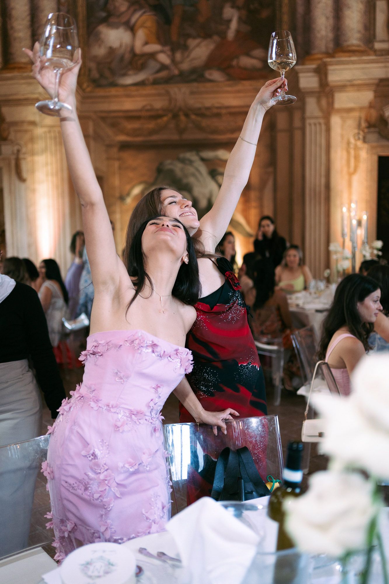A person in a pink dress raises two wine glasses in celebration at an elegant indoor reception venue.