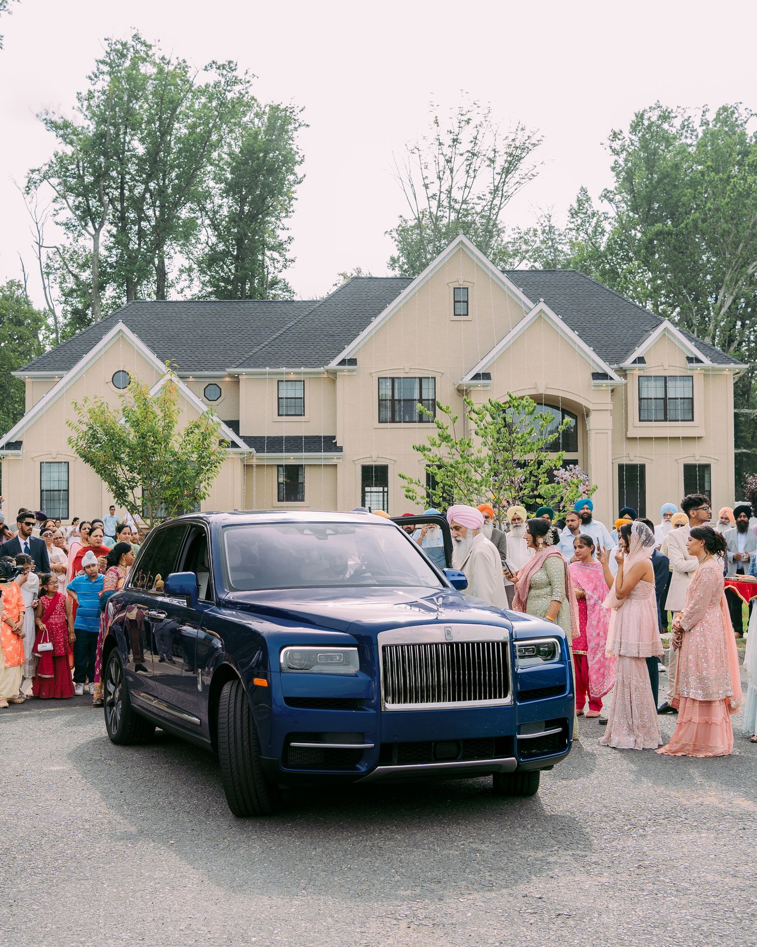 A luxury blue Rolls-Royce Cullinan SUV parked in front of a beige suburban house with guests gathered around.