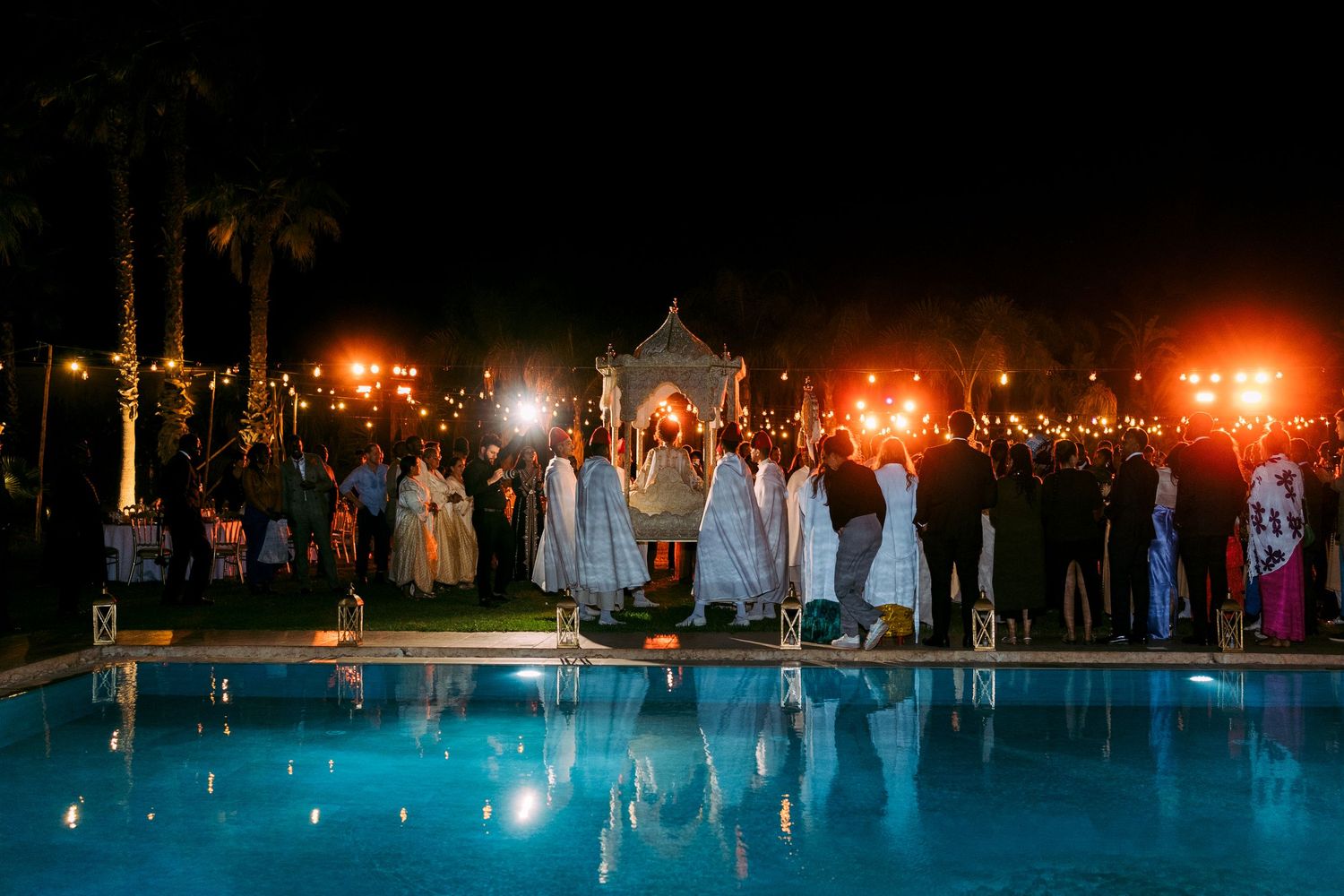 Wedding guests gathered around an illuminated pool during an evening outdoor reception.
