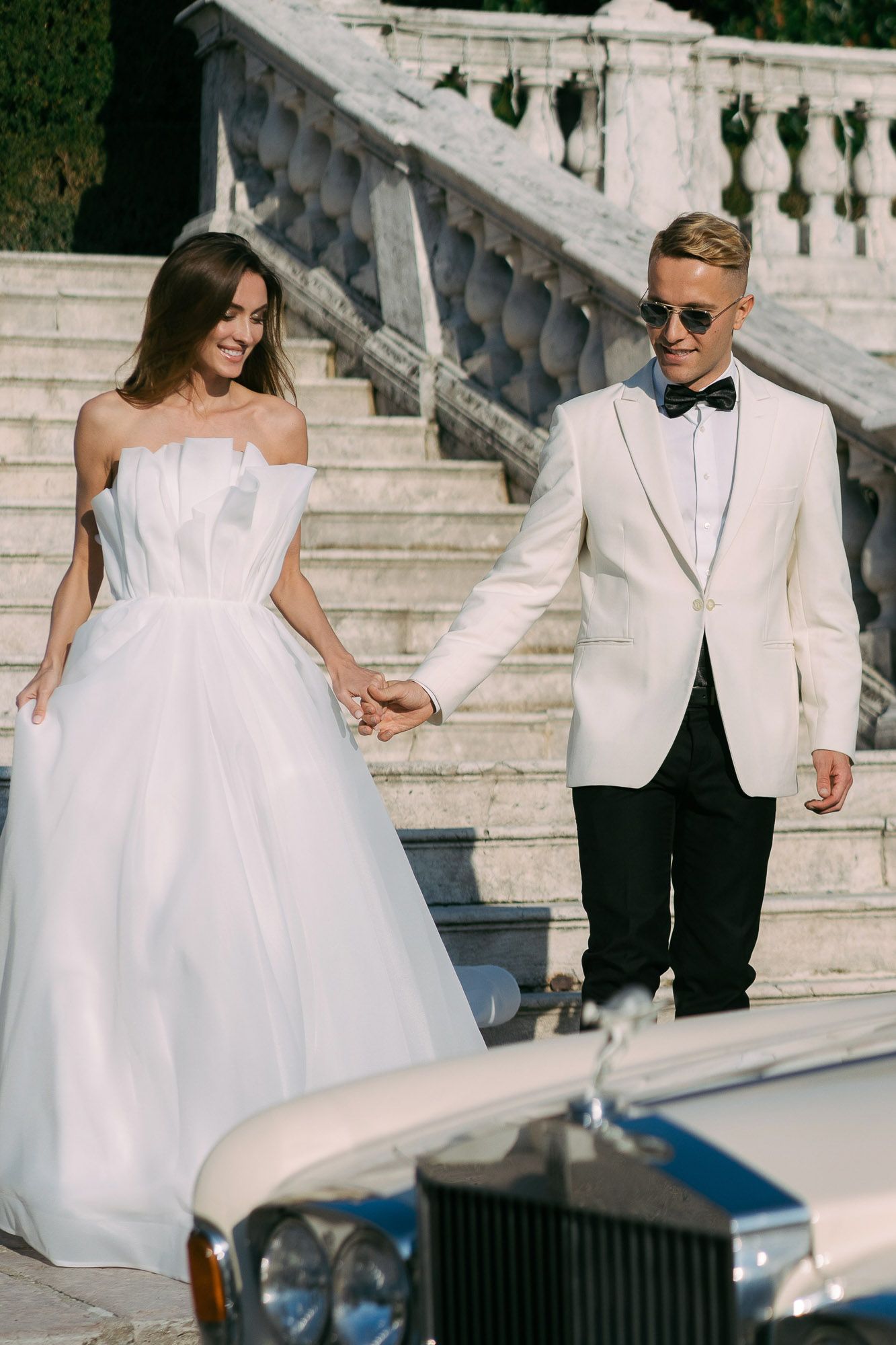 Couple in elegant white wedding attire descending stone steps beside a vintage Rolls Royce luxury car.