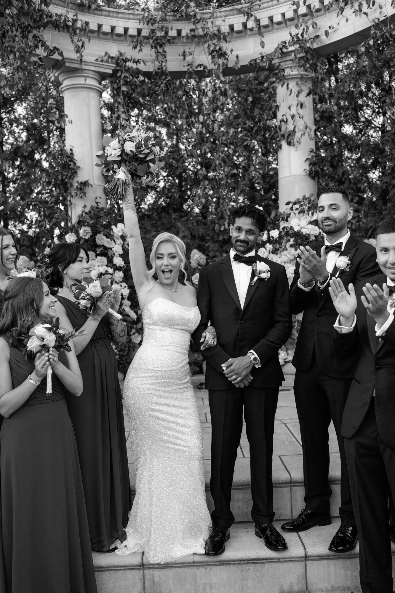 Wedding party in black and white formal attire poses together on steps with joyful expressions and laughter.