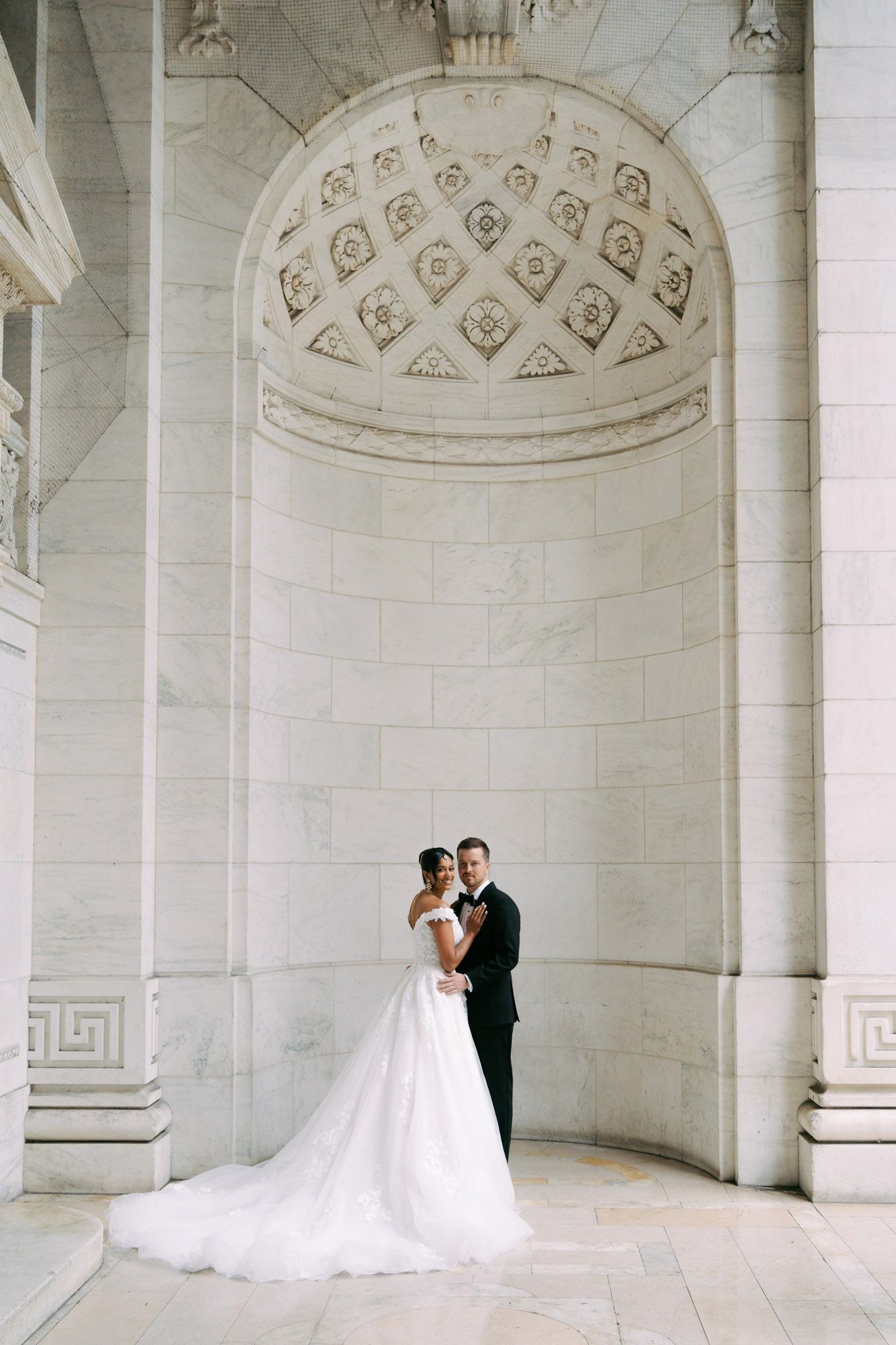 Elegant couple stands between ornate marble columns in a classical architectural setting with arched details.