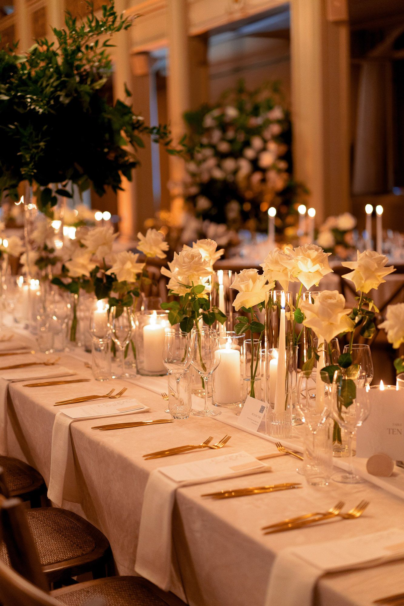 Elegant wedding reception tables with white linens, candlelight, and floral centerpieces in a warm-lit venue.
