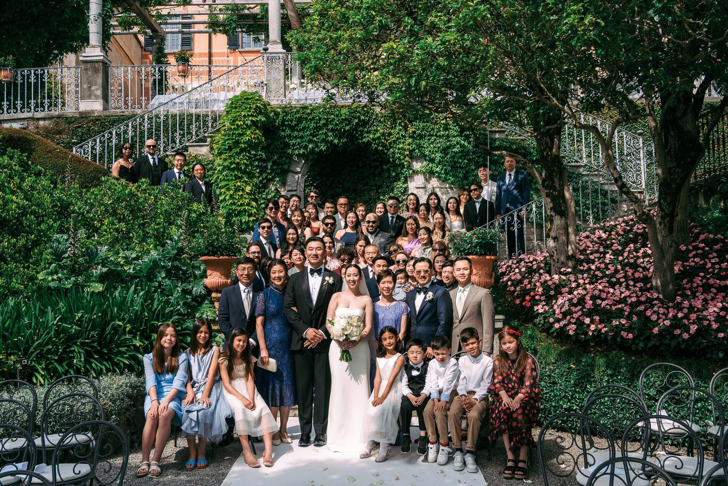 Large group of wedding guests pose for photos in a lush garden setting with brick buildings in the background.