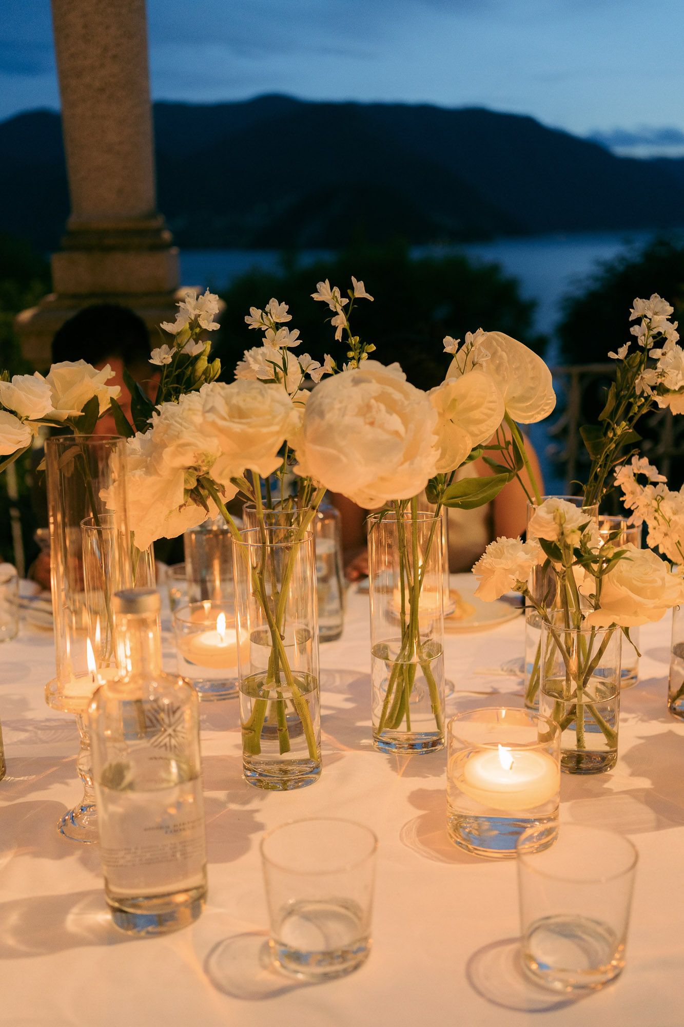 White roses and small flowers in glass vases with glowing candles create an elegant evening table setting by the water.