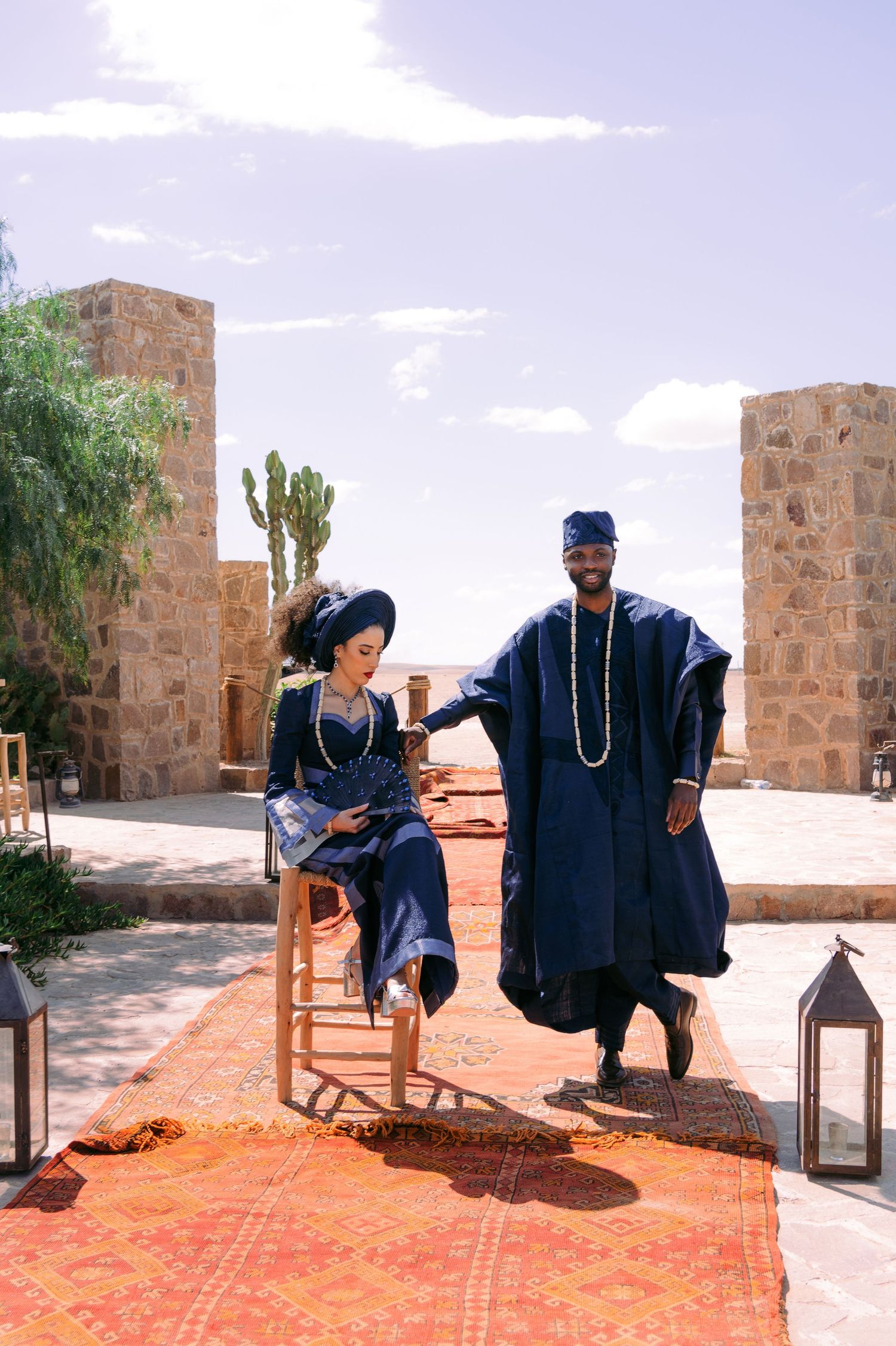 Two figures in traditional blue robes sit on chairs on an outdoor tiled patio near stone walls in a desert setting.