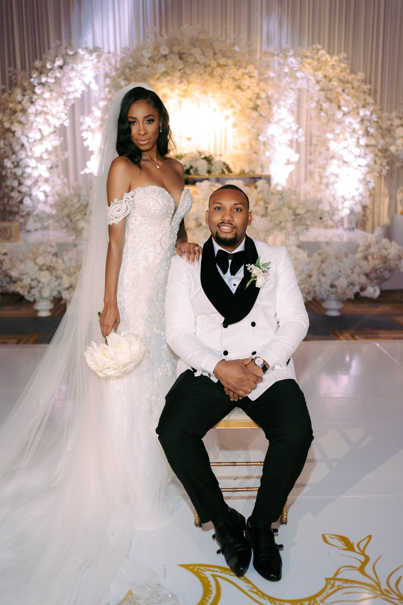 A bride and groom pose together on an elegant white and gold stage with floral decorations at their wedding reception.