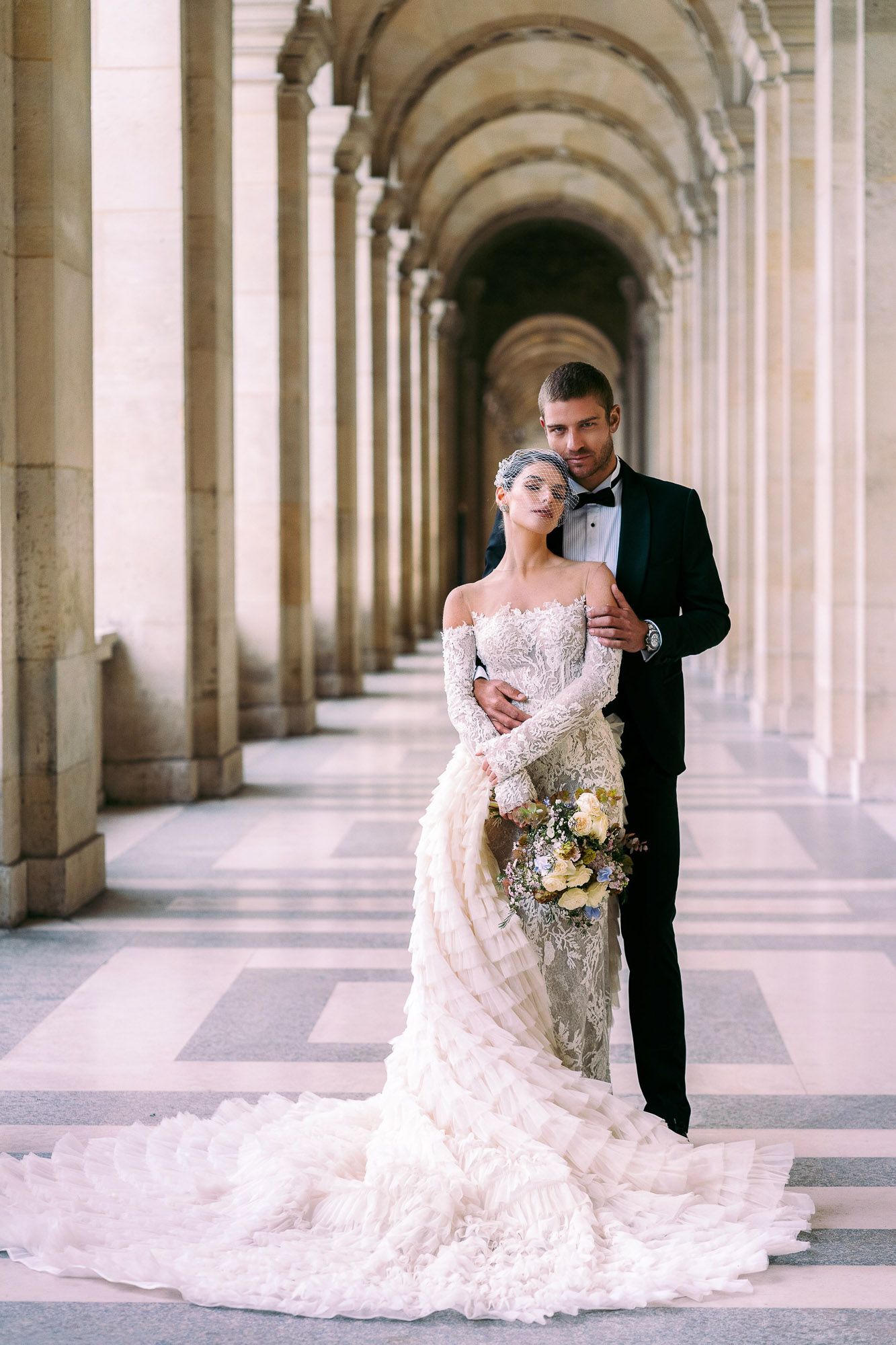 A bride and groom embrace in an elegant colonnade hallway with arched architecture and soft natural lighting.
