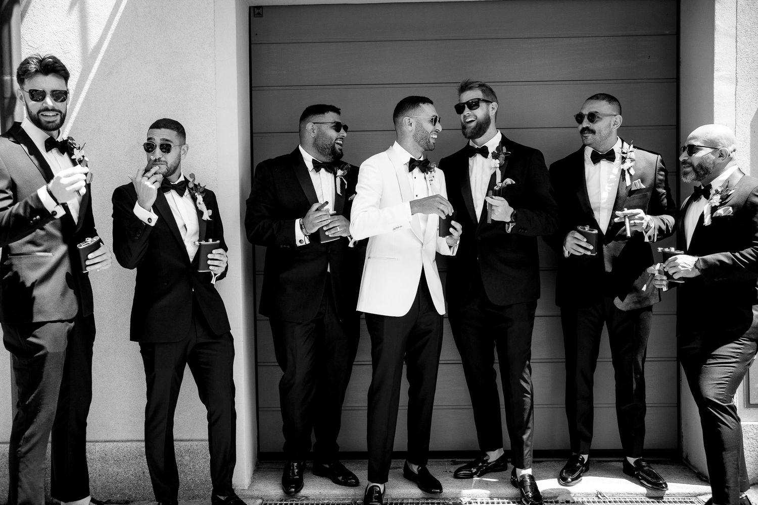 A group of groomsmen in formal attire share a candid moment laughing together in black and white photography.