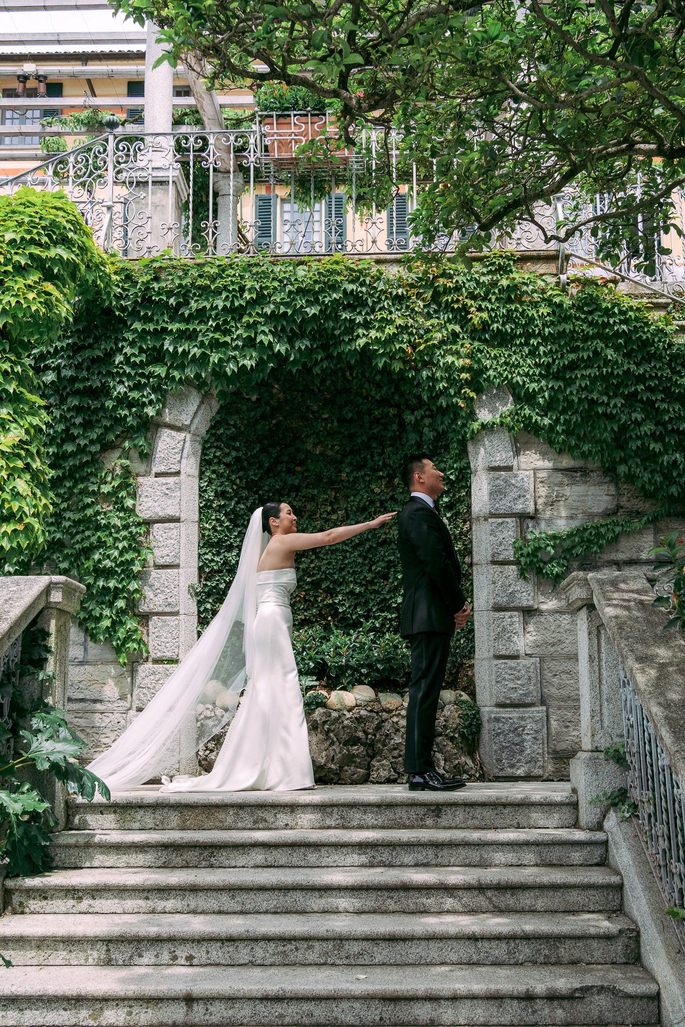 A romantic scene of a couple in wedding attire sitting and posing on stone steps surrounded by lush green ivy.