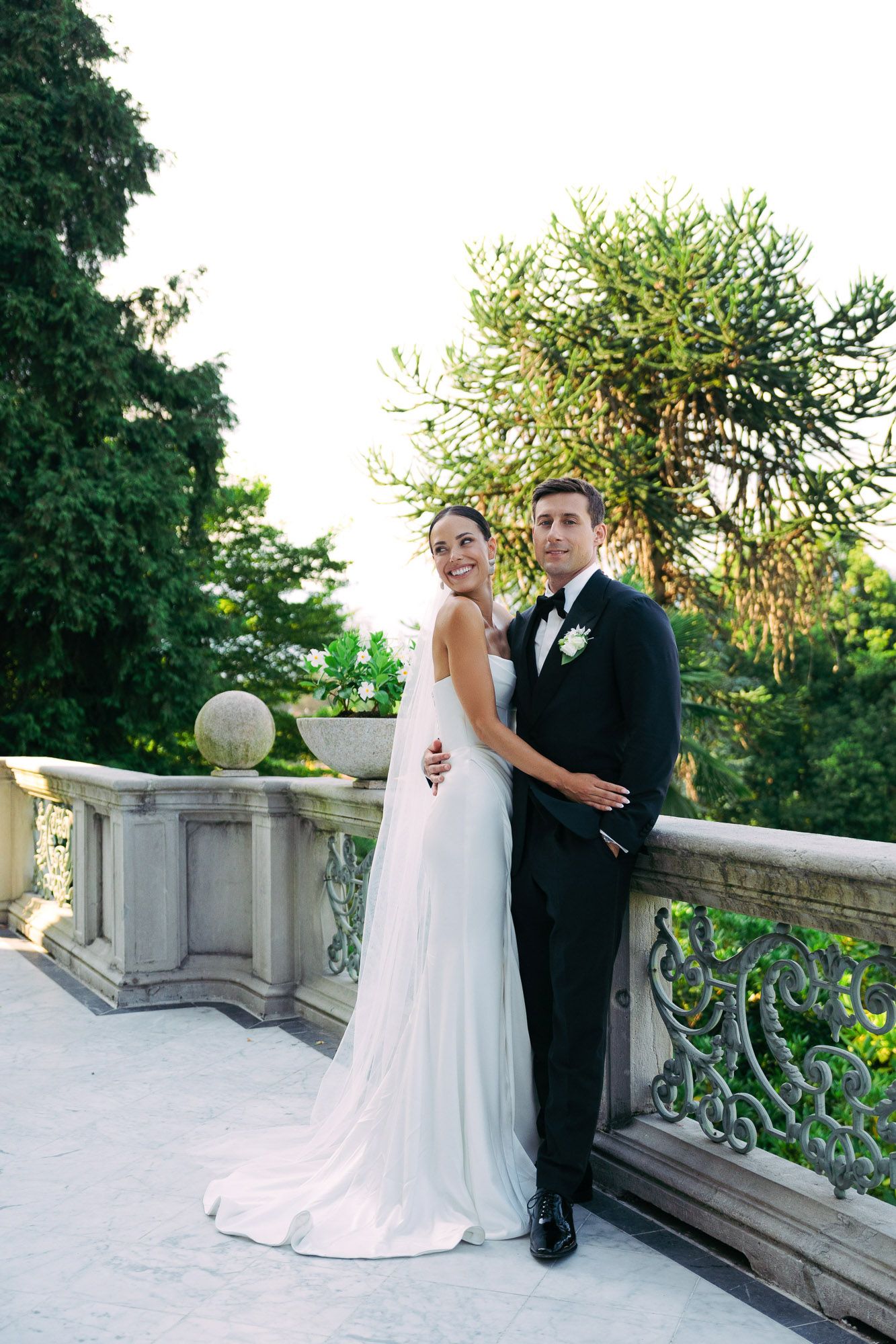 Elegant wedding couple poses on stone balcony at sunset with lush greenery in background.