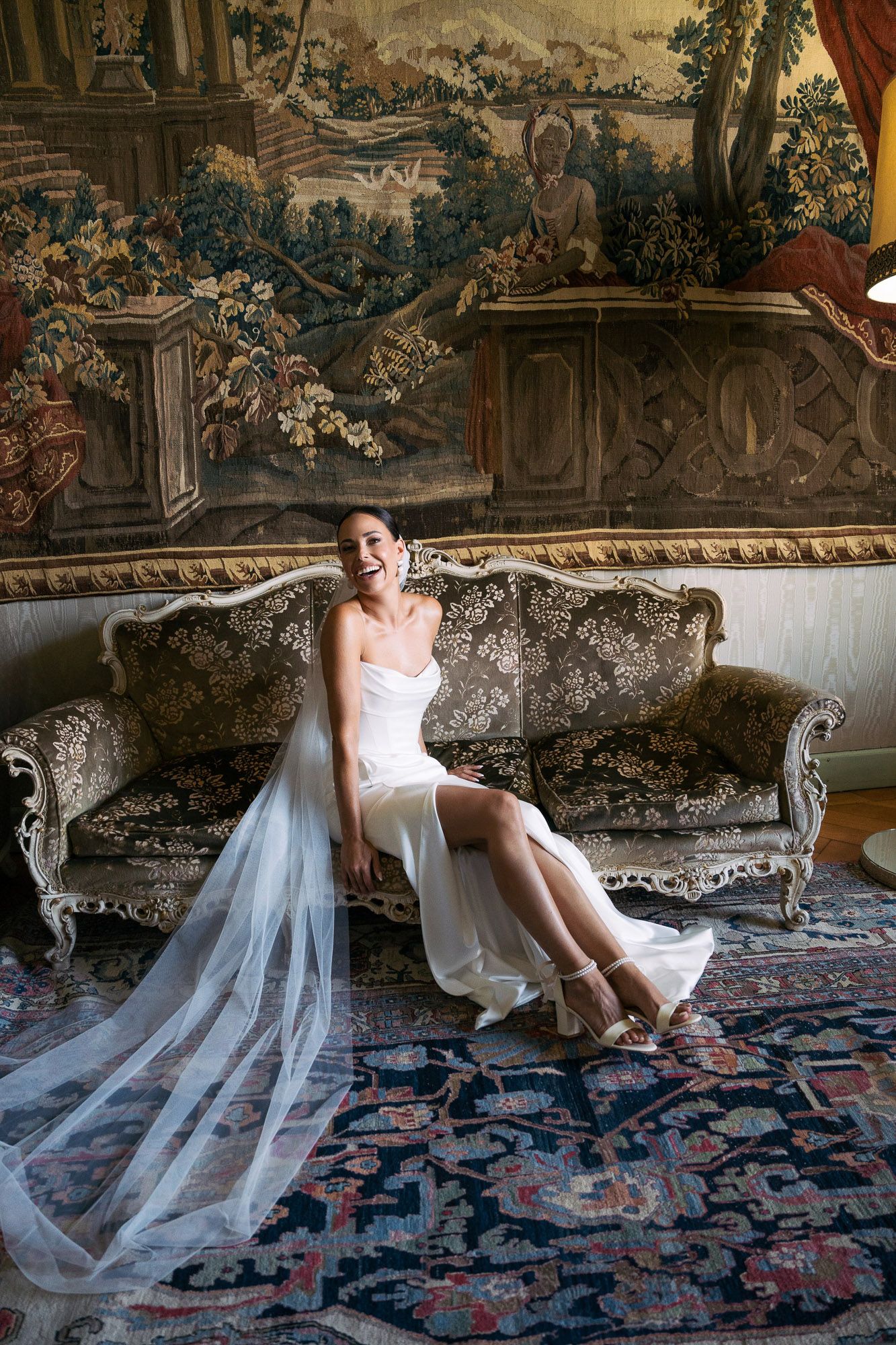 A bride in a flowing white dress reclines on an antique chaise lounge against ornate tapestry walls.