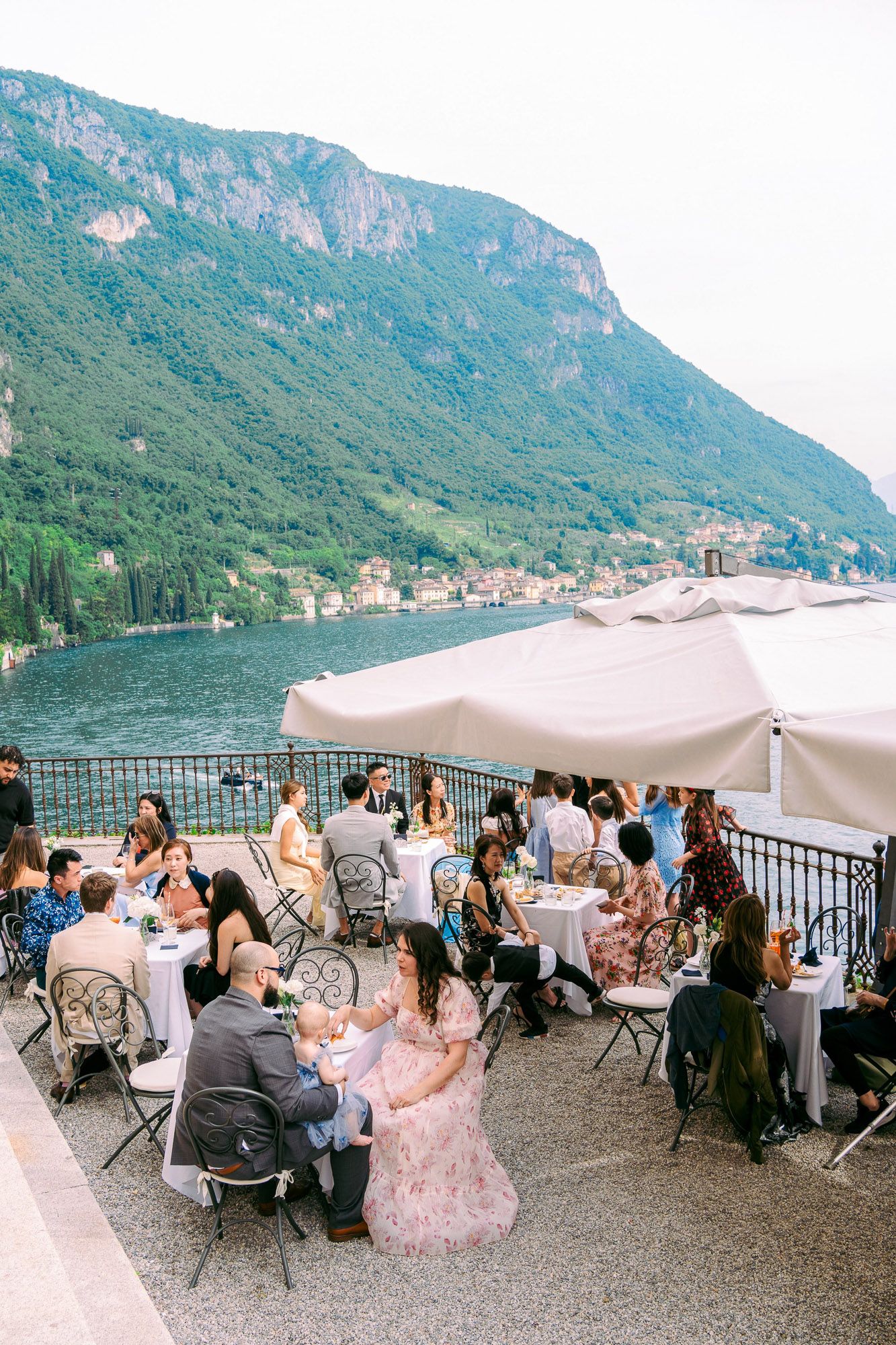 Wedding guests dine at outdoor tables with white umbrellas overlooking a scenic mountain lake in Italy.