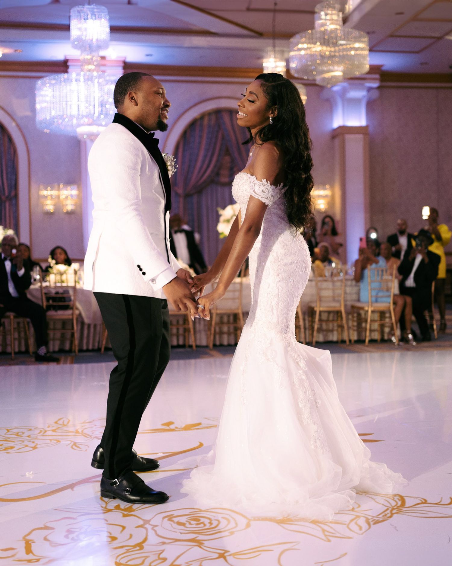 A couple shares their first dance at an elegant wedding reception with crystal chandeliers and ornate floor designs.