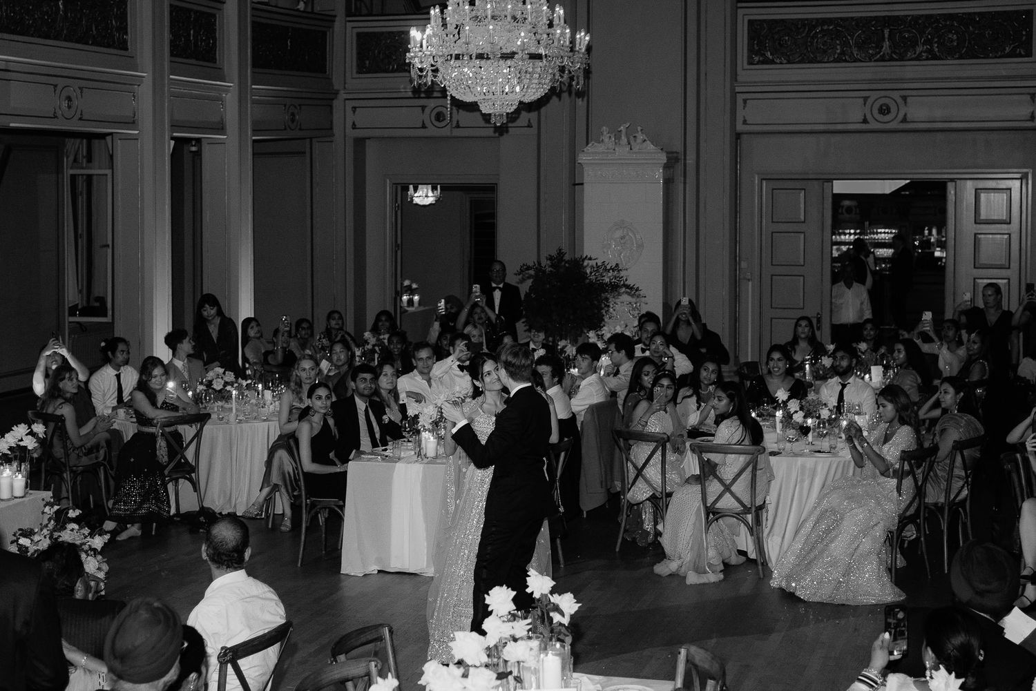 Black and white photo of a wedding reception with guests seated at long tables under ornate chandeliers.