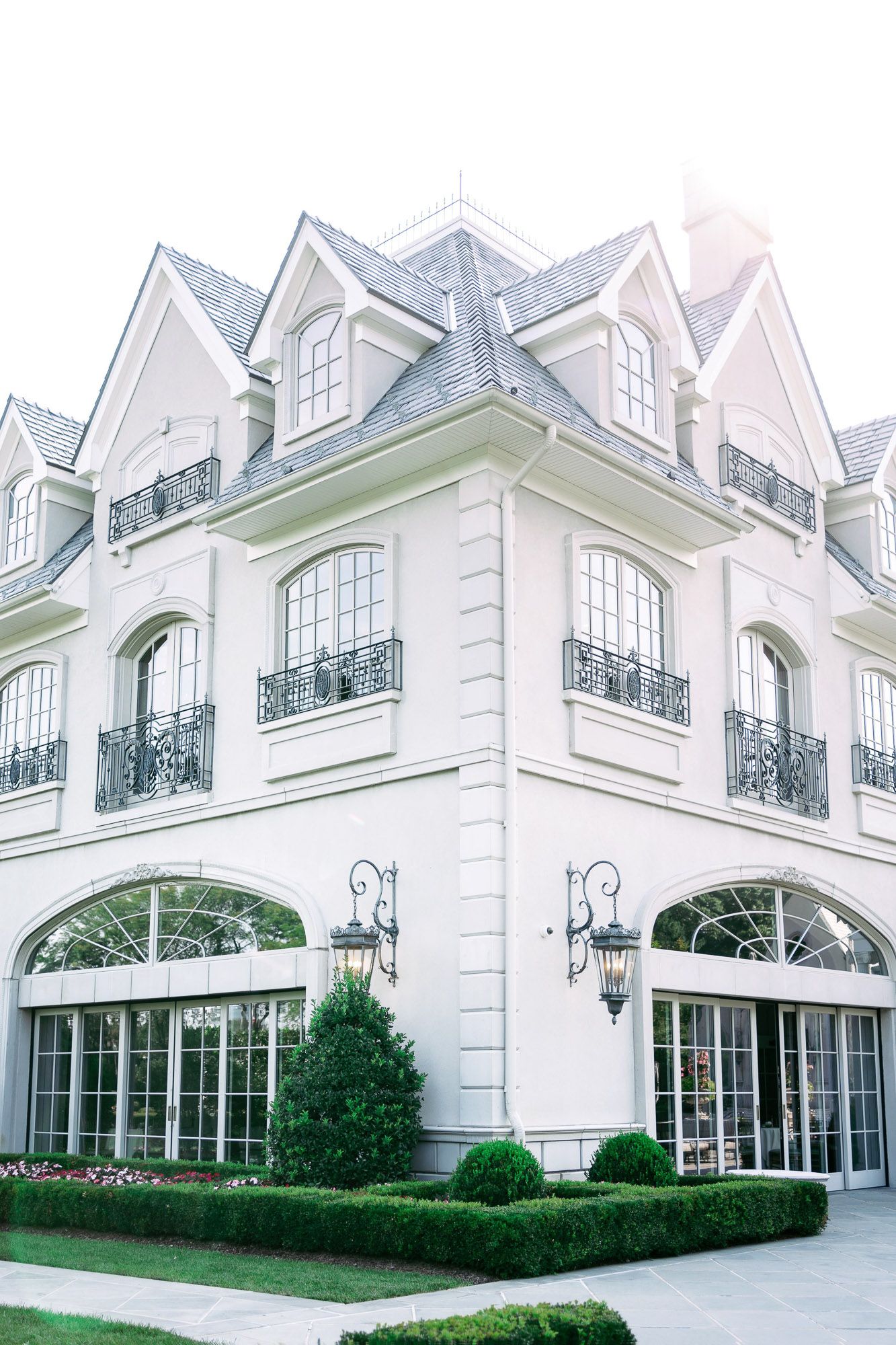 Elegant white French-style mansion with arched windows, wrought iron balconies, and manicured hedges along the facade.