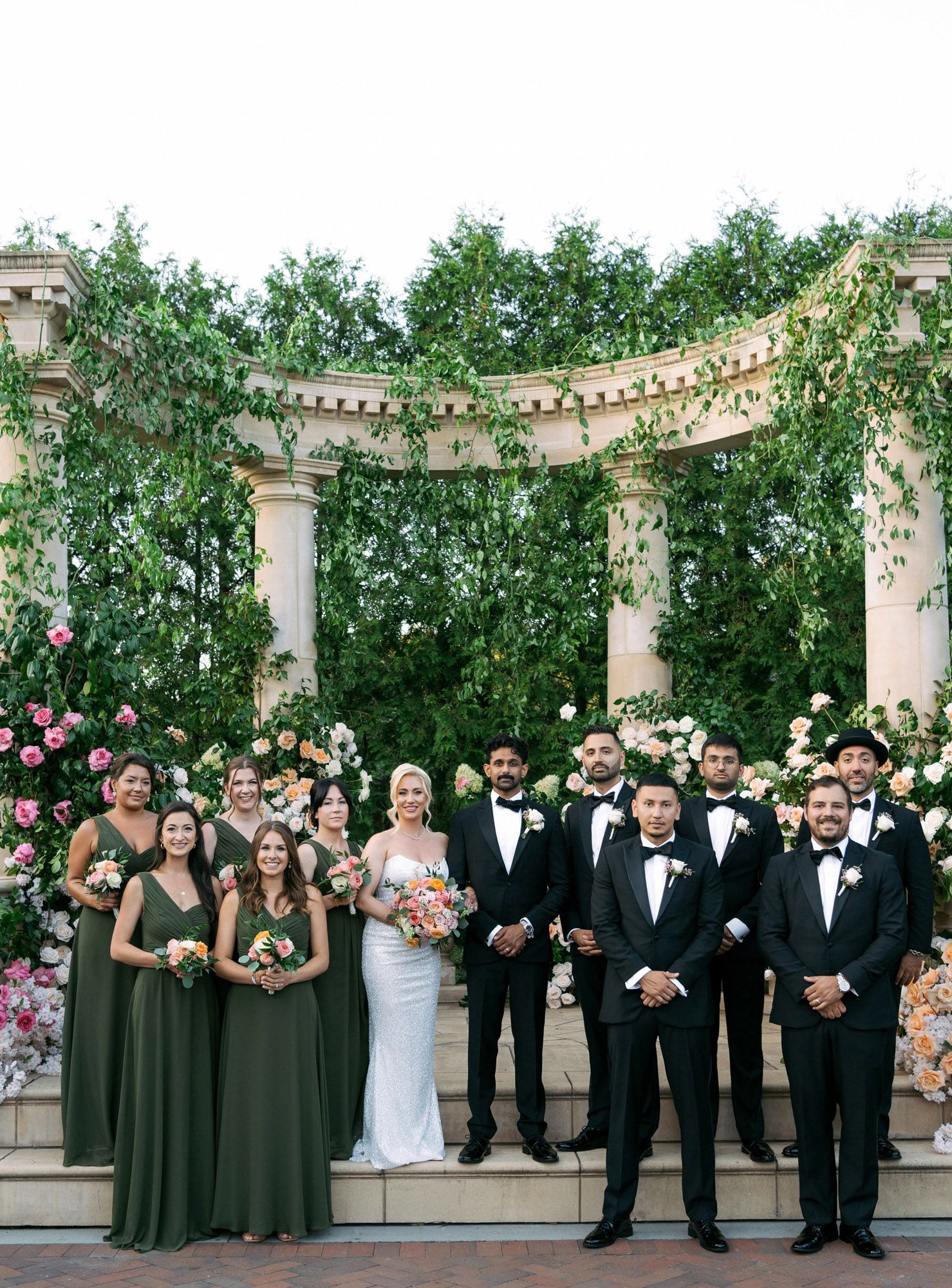 Wedding party in black tuxedos and dark green dresses pose together under a floral-decorated pergola.