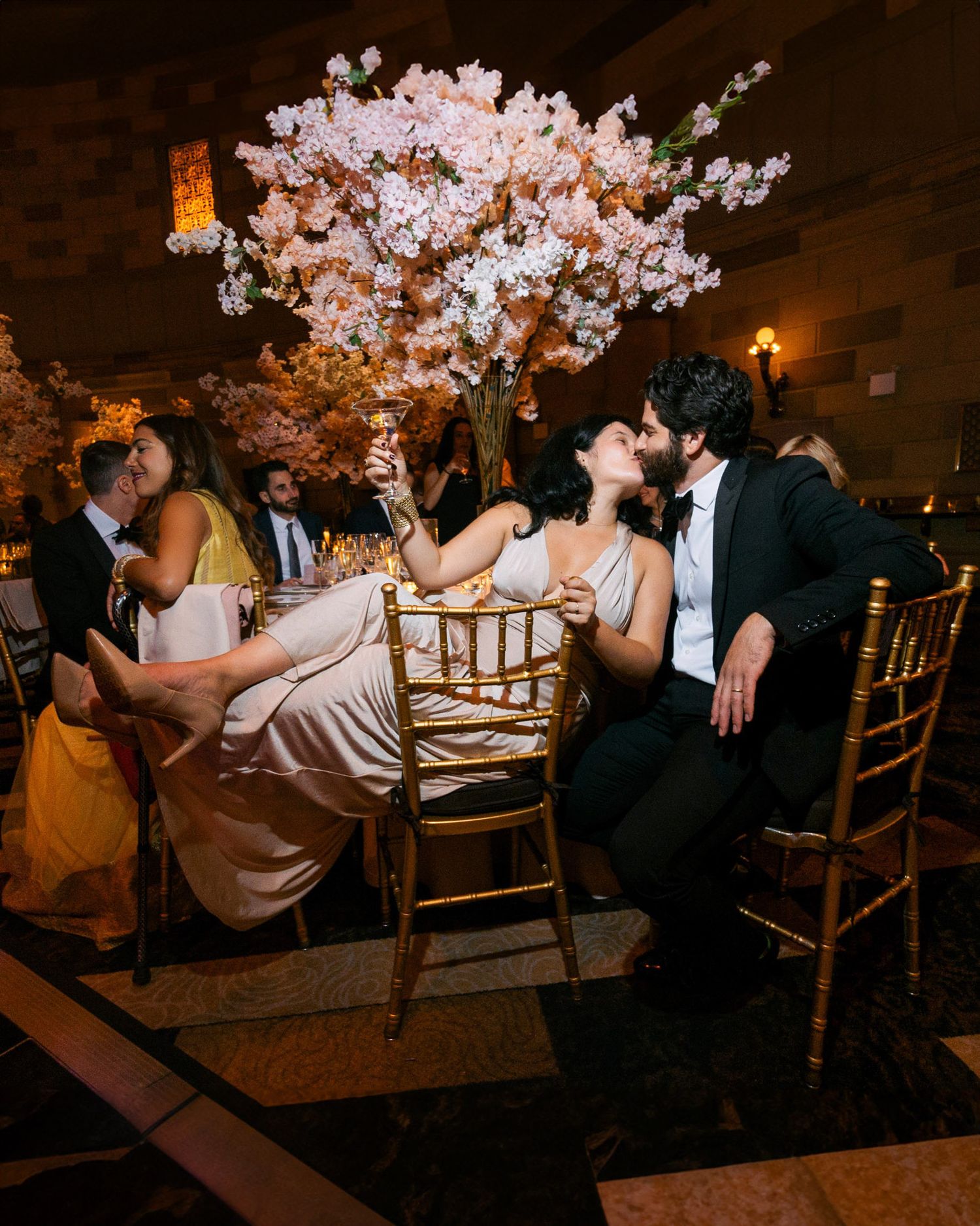 Wedding reception guests sit at elegantly decorated tables with tall white floral centerpieces in a dimly lit ballroom.