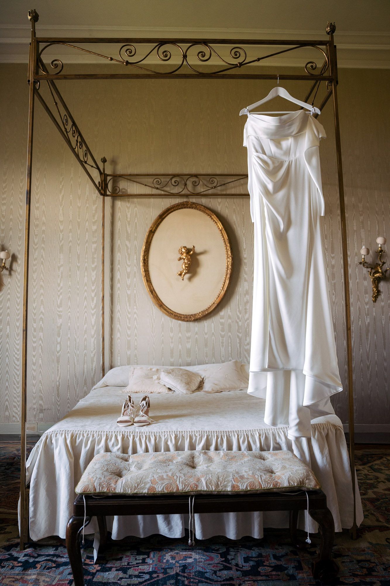 A vintage wrought iron canopy bed with white linens and ruffled bedskirt sits in an elegant bedroom with floral wallpaper.