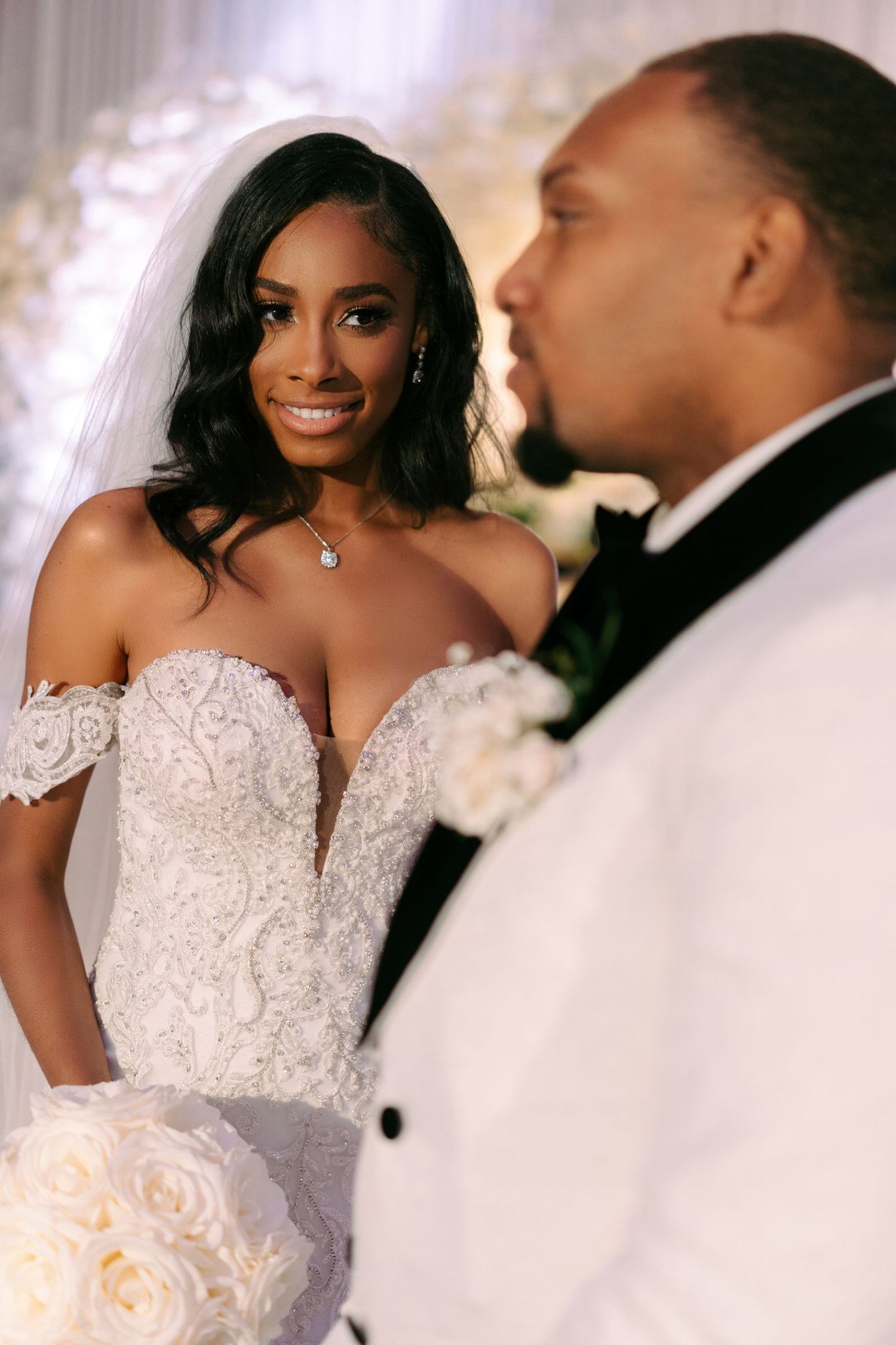 Elegant bride in lace off-shoulder wedding gown smiles while holding white rose bouquet next to groom in white tuxedo jacket.