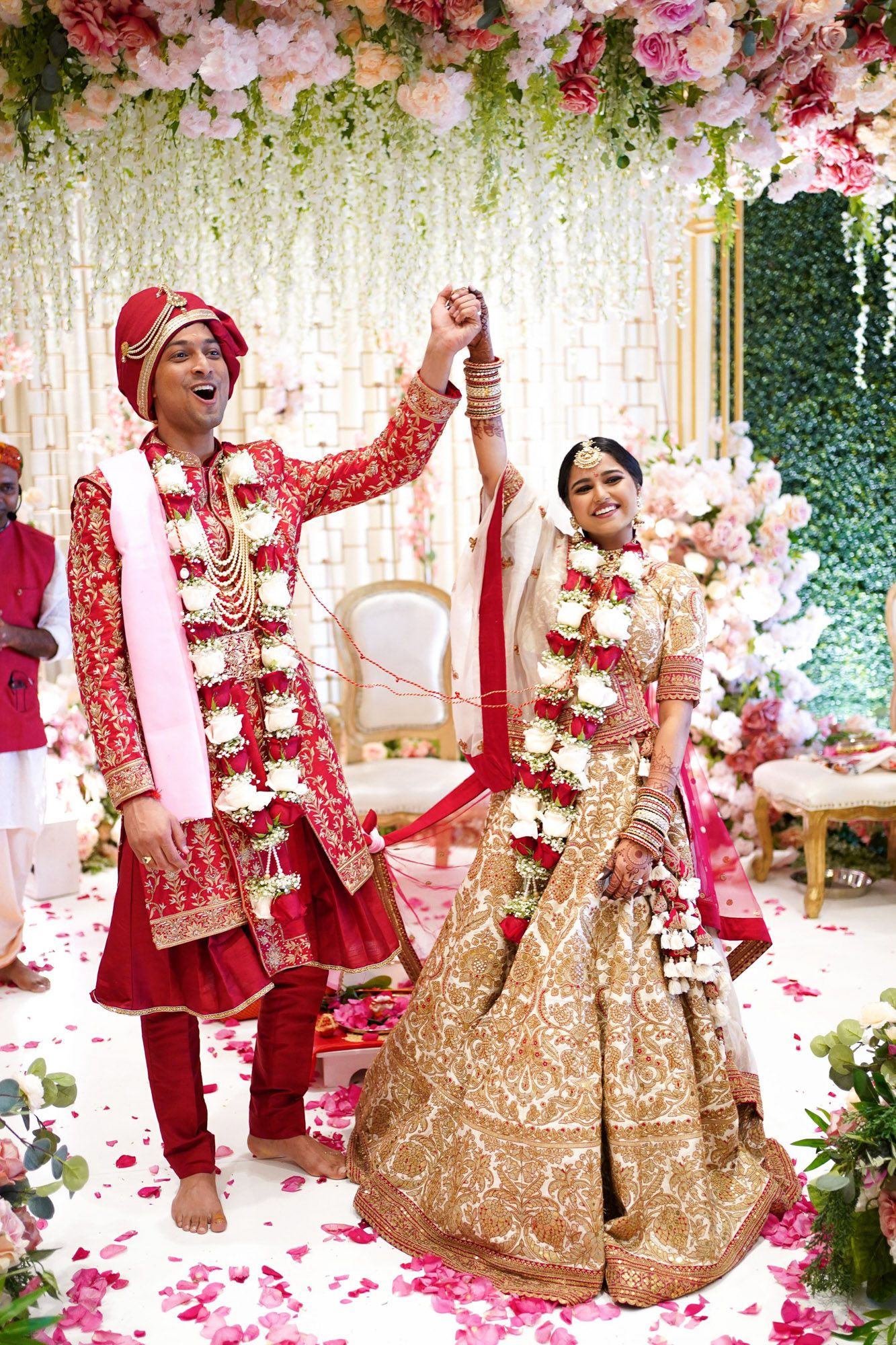 Traditional Indian wedding ceremony celebration with red and gold attire amidst pink floral decorations and white drapery.