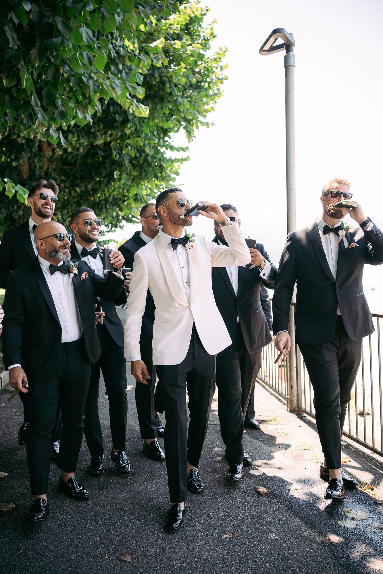 A sequence of groomsmen in black tuxedos and bow ties striking fun poses together outdoors near greenery.
