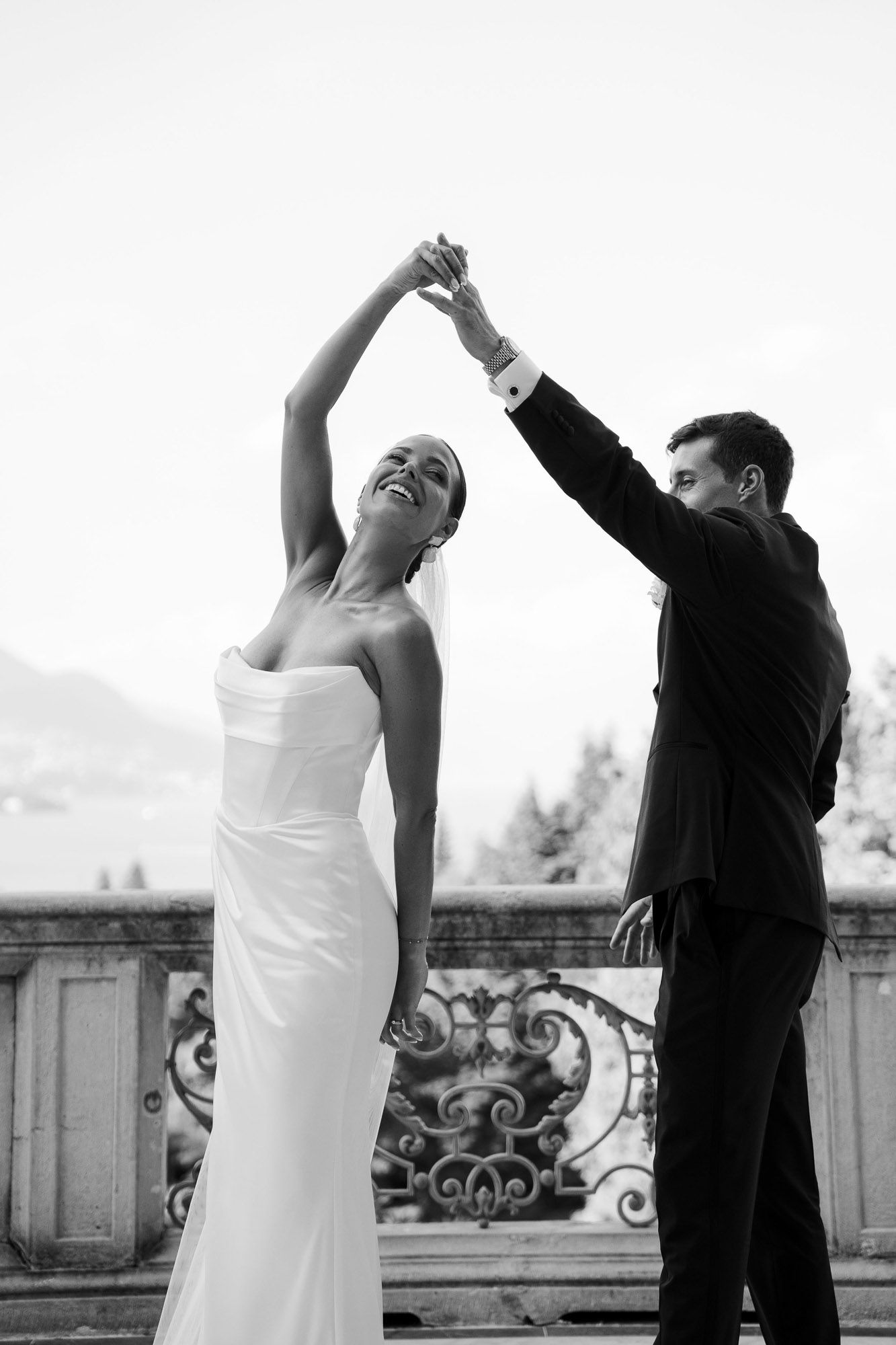Black and white photo of a couple dancing together on a balcony with ornate railing, captured in an elegant pose.