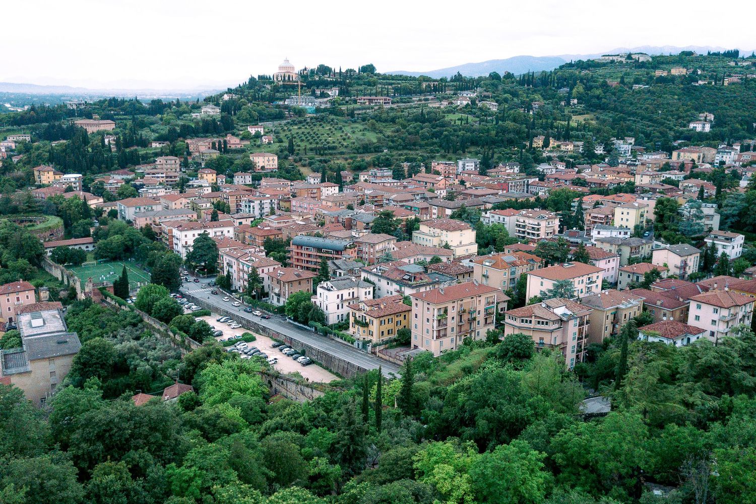 Panoramic view of a hillside Mediterranean town with terracotta roofs nestled among lush green trees.