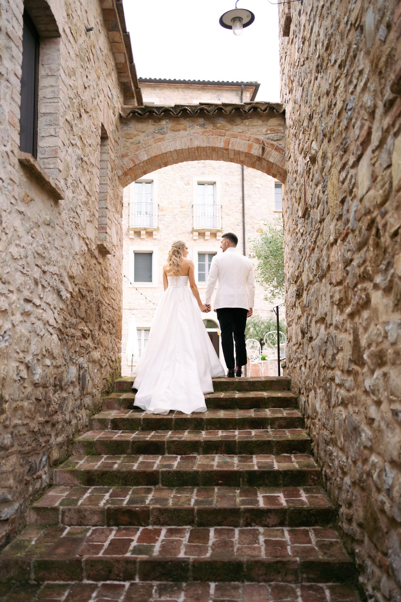 A couple ascends stone steps in a rustic Mediterranean courtyard with brick and stone walls.