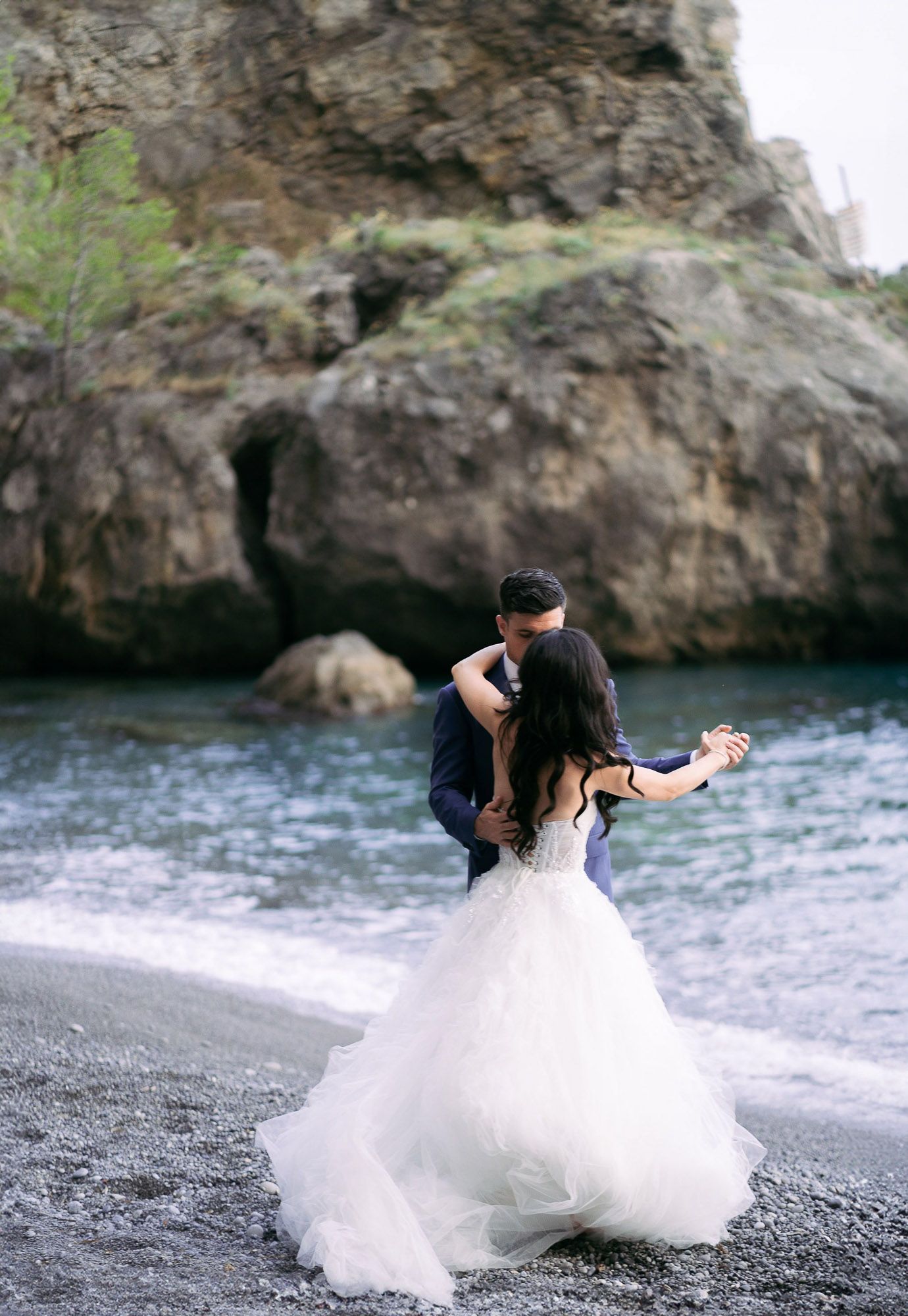 A couple in wedding attire dances romantically on a black sand beach with dramatic cliffs in the background. wedding photographer Italy