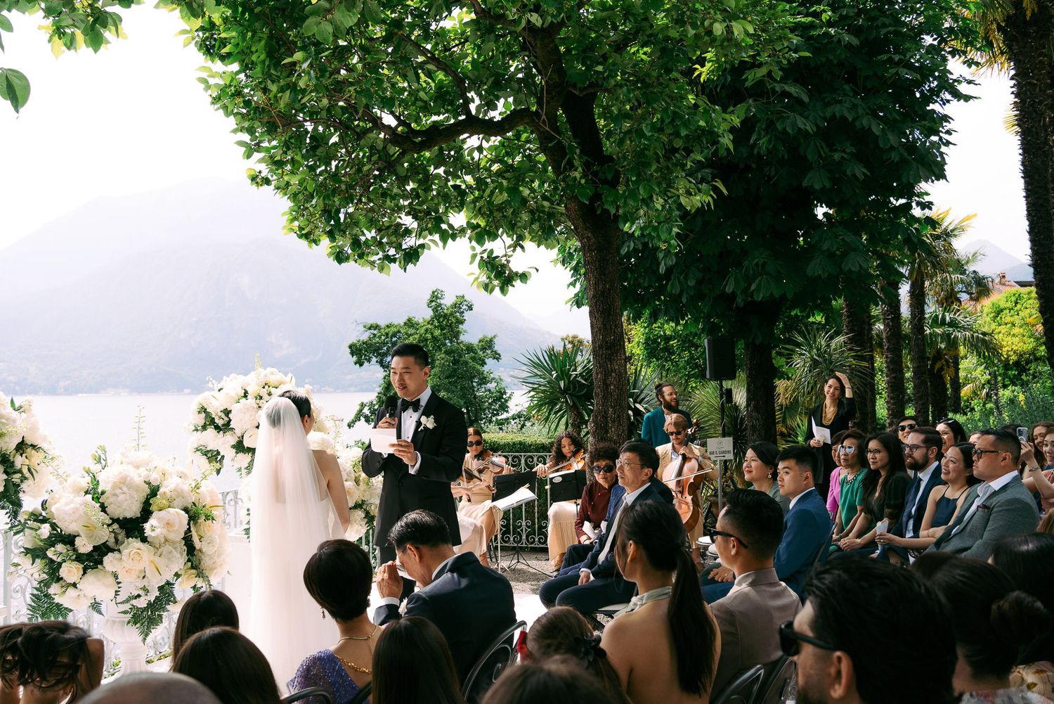 A panoramic view of an outdoor wedding ceremony under trees with guests seated on white chairs.