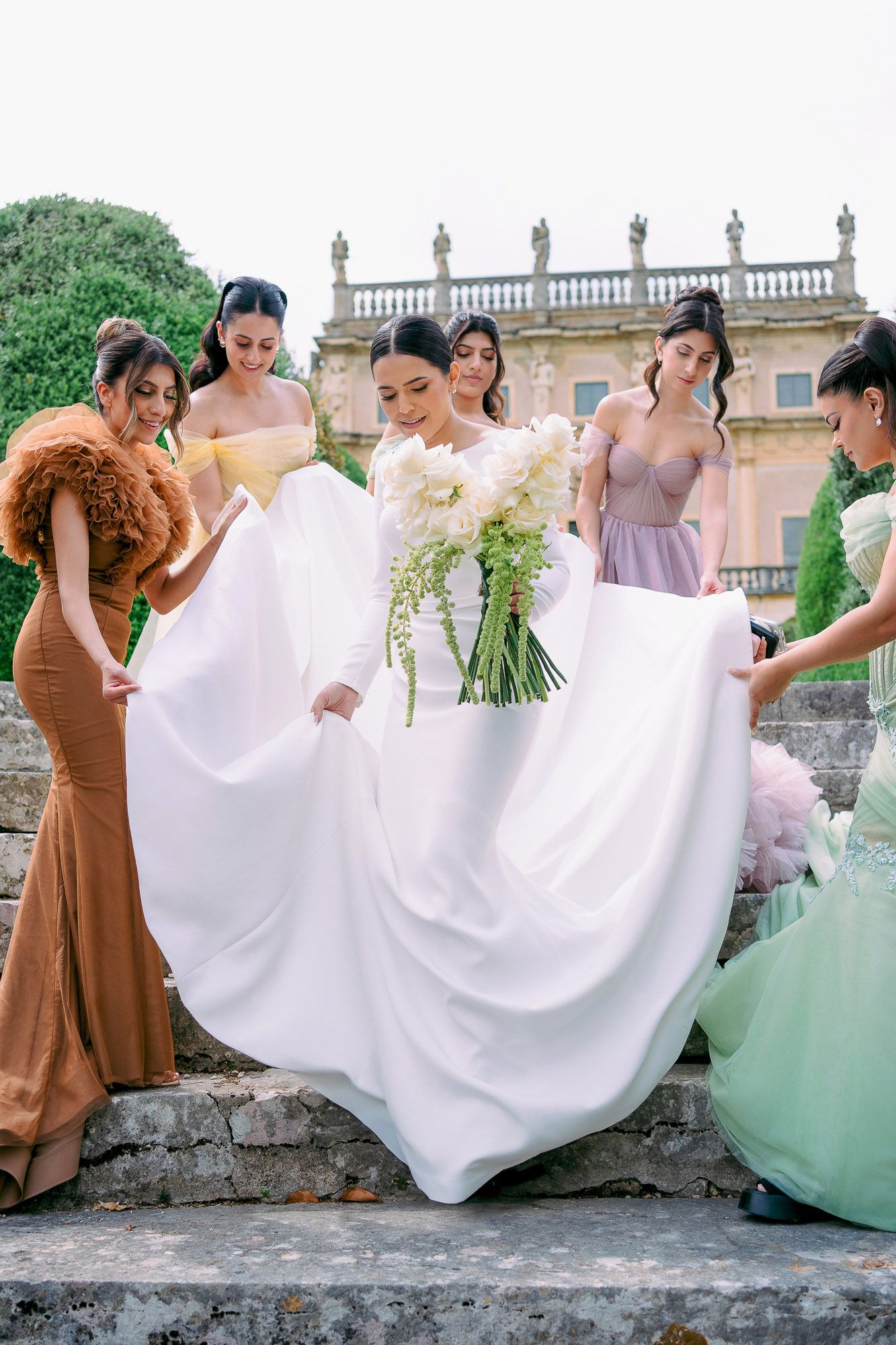 Bridesmaids in mint green and brown dresses help adjust a white wedding gown train on stone steps outdoors.