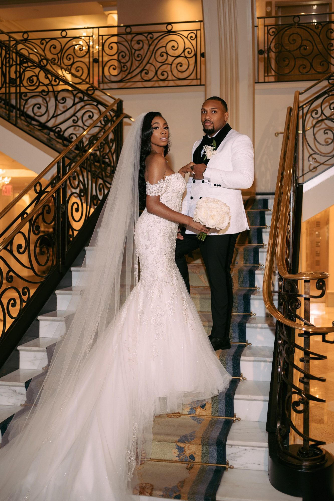 A couple in wedding attire poses on an ornate staircase with wrought iron railings during their photoshoot.